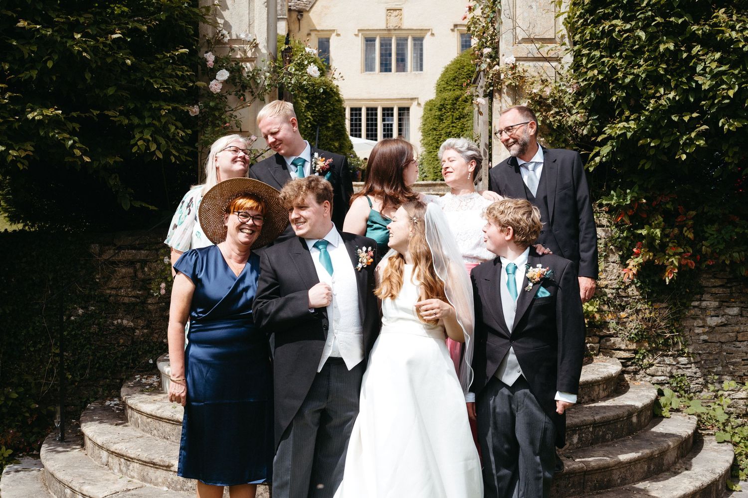 Wedding party gathers on stone steps of elegant old building for formal group photo in sunlight.