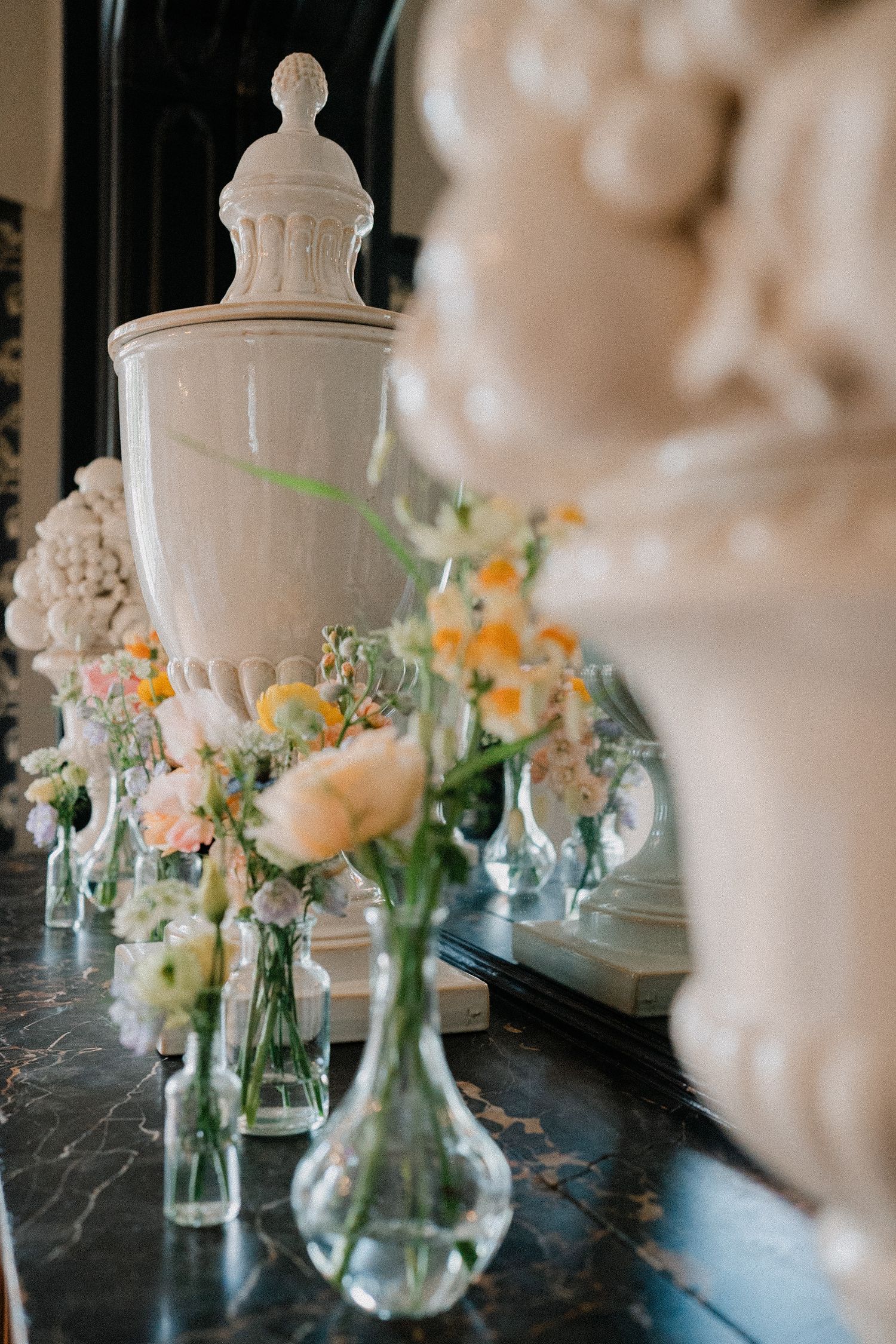 White decorative urn with delicate spring flower arrangements in glass vases on dark marble surface.