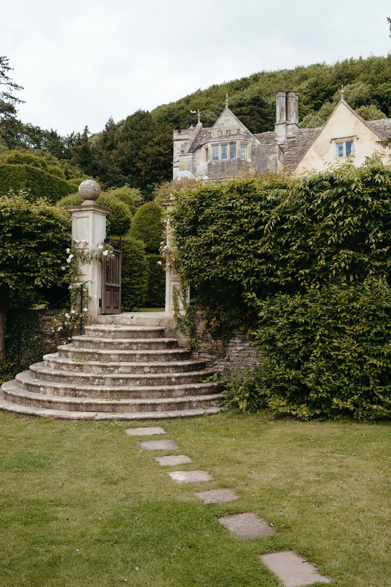 Grand stone staircase with ornate pillars leading to an English manor house surrounded by manicured hedges.