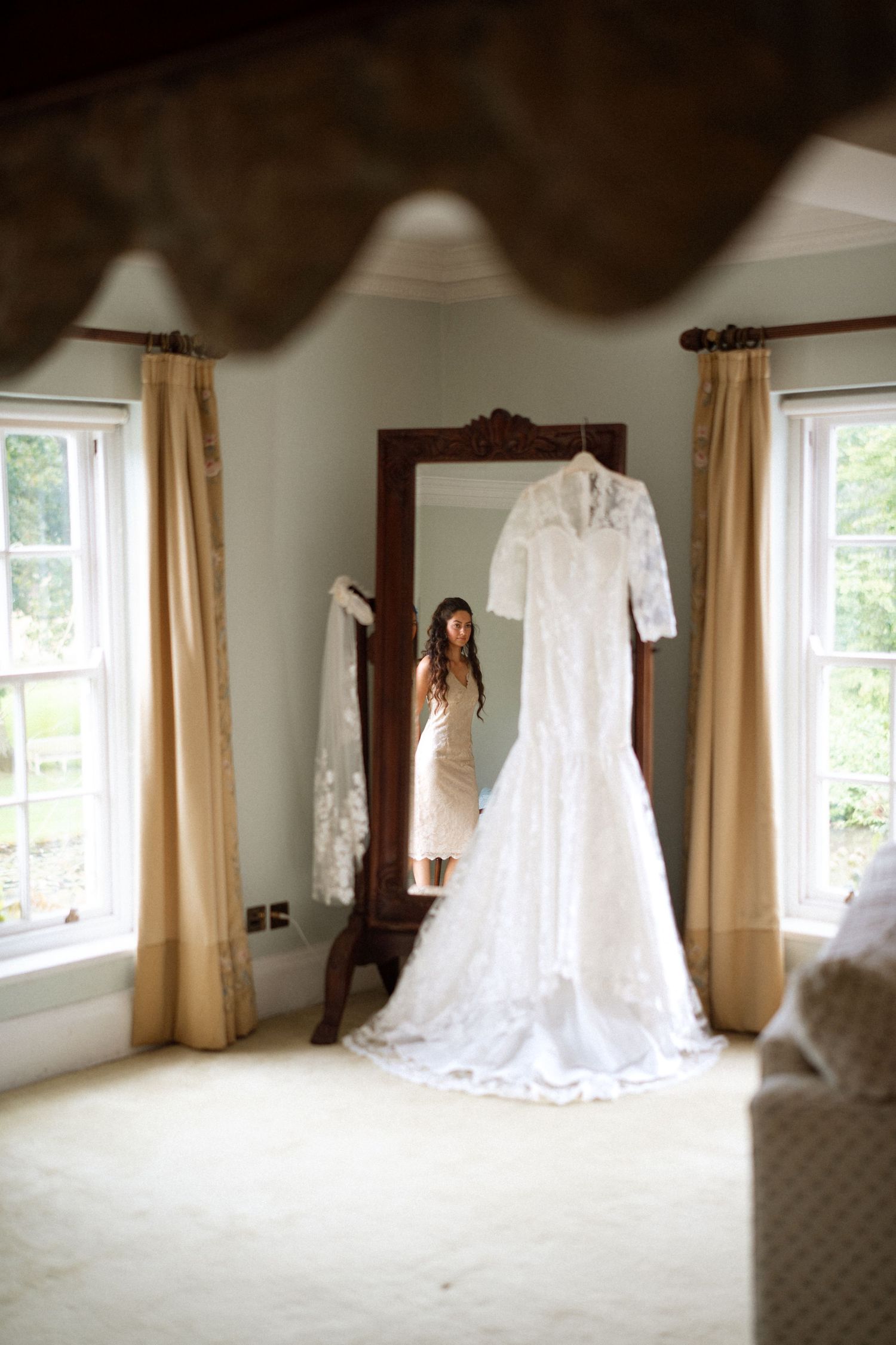 A white lace wedding dress hanging in a sunlit room with cream curtains.
