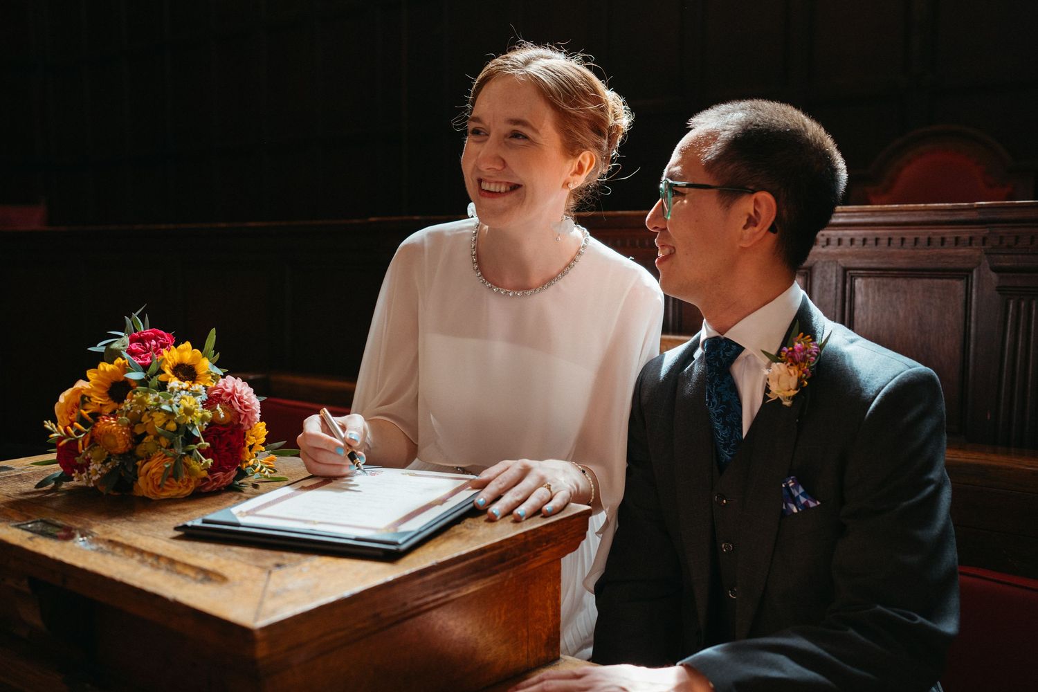 A couple shares a joyful moment while signing documents at a wooden desk.