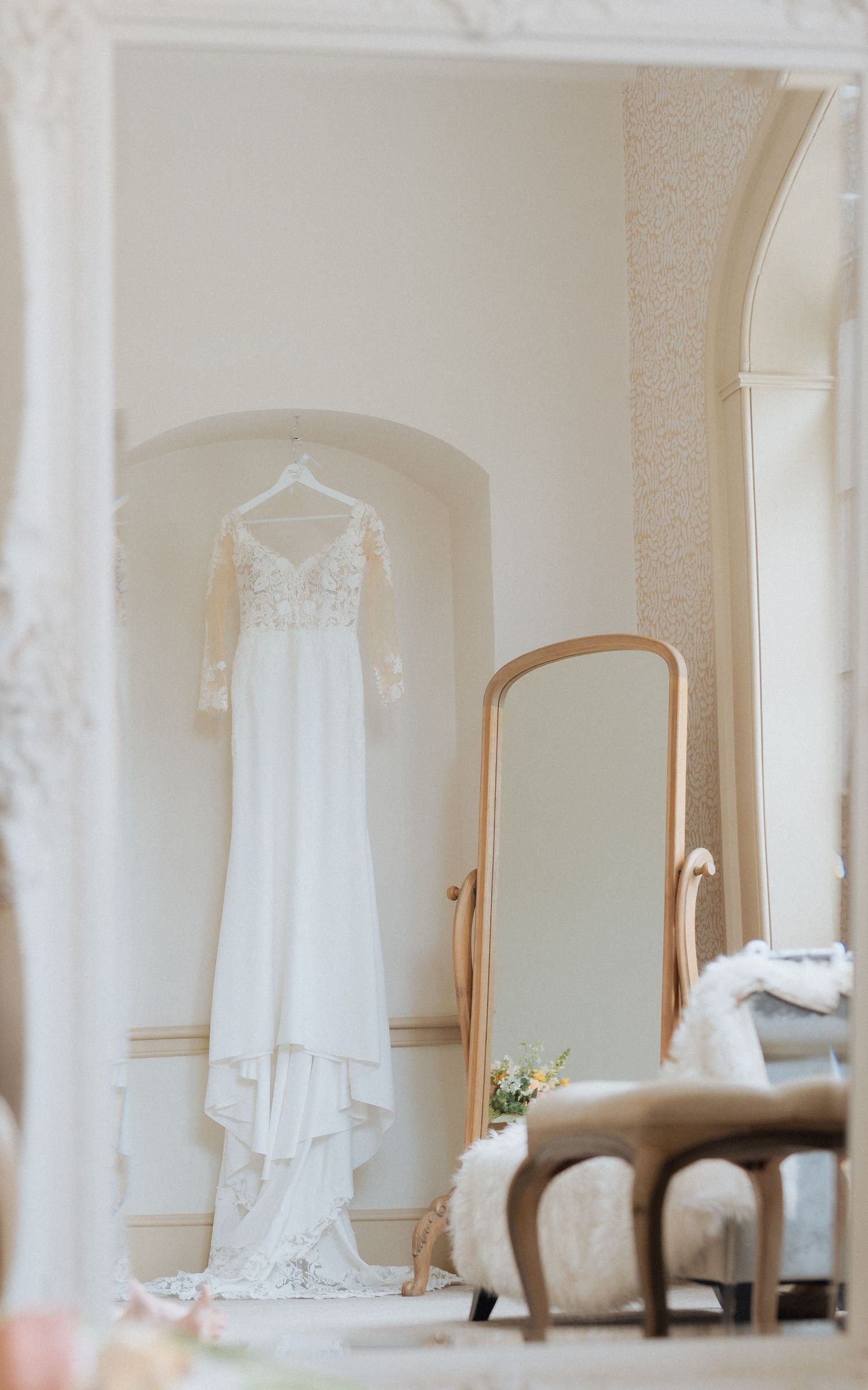 A white lace wedding dress hangs in an elegant room with vintage furniture and a standing mirror. Wedding at Nonsuch Mansion