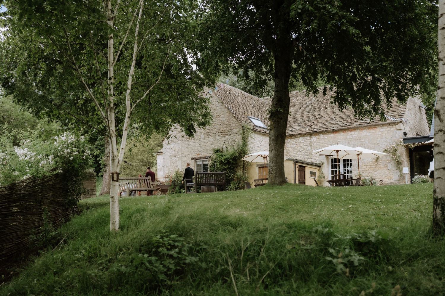 Rustic stone barn nestled into a green hillside with trees and natural landscaping creating a peaceful countryside scene.