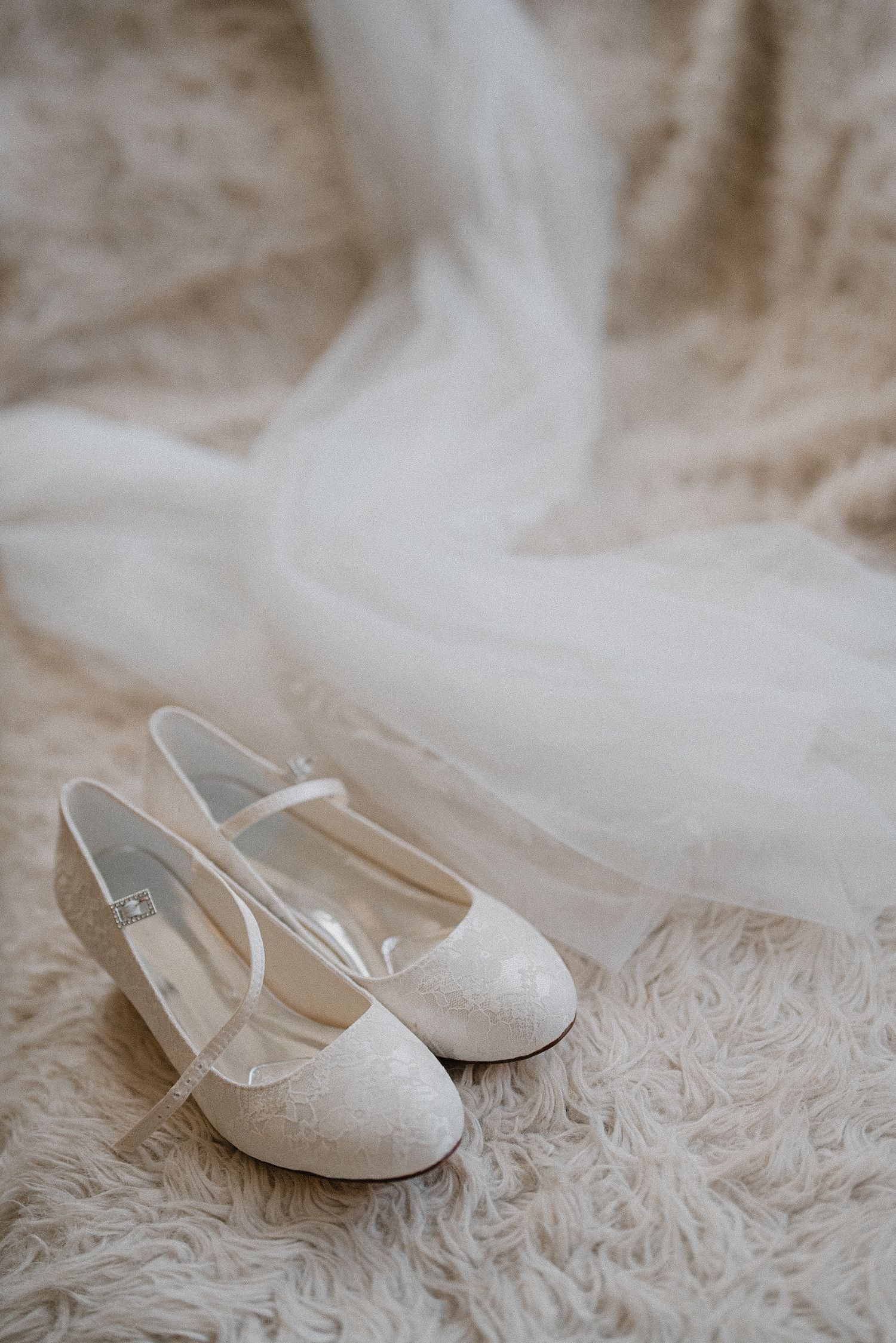 White lace bridal flats and delicate tulle veil displayed on a soft furry carpet for a wedding day.