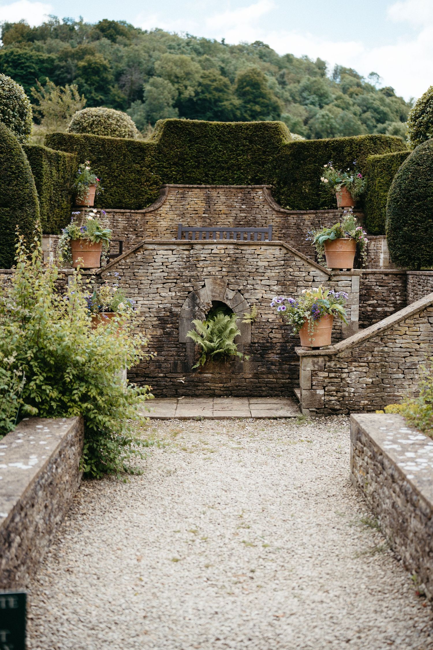 Stone pathway leading to an elegant garden wall with ornamental topiaries and potted plants at a historic estate.