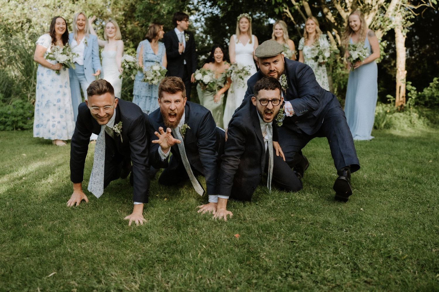 A wedding party poses playfully on a green lawn with people in blue dresses and dark suits creating a fun group photo.