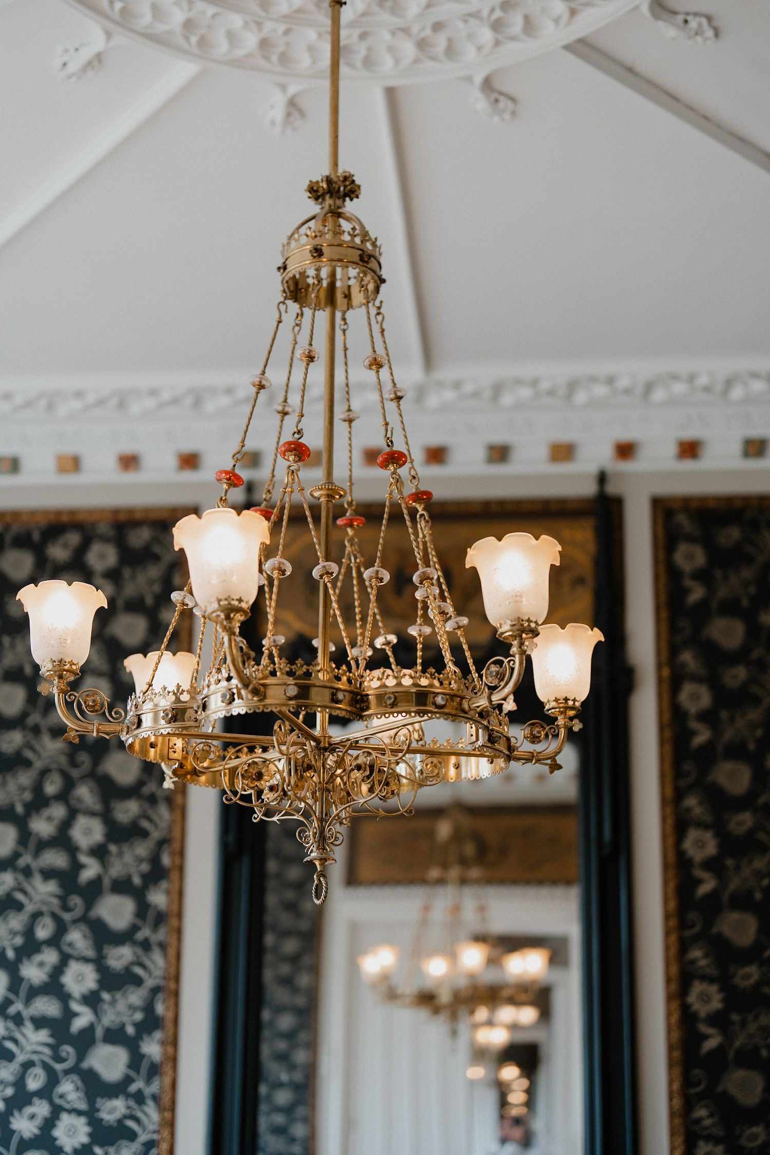 Elegant vintage brass chandelier with frosted glass shades hangs from ornate white ceiling with decorative molding.
