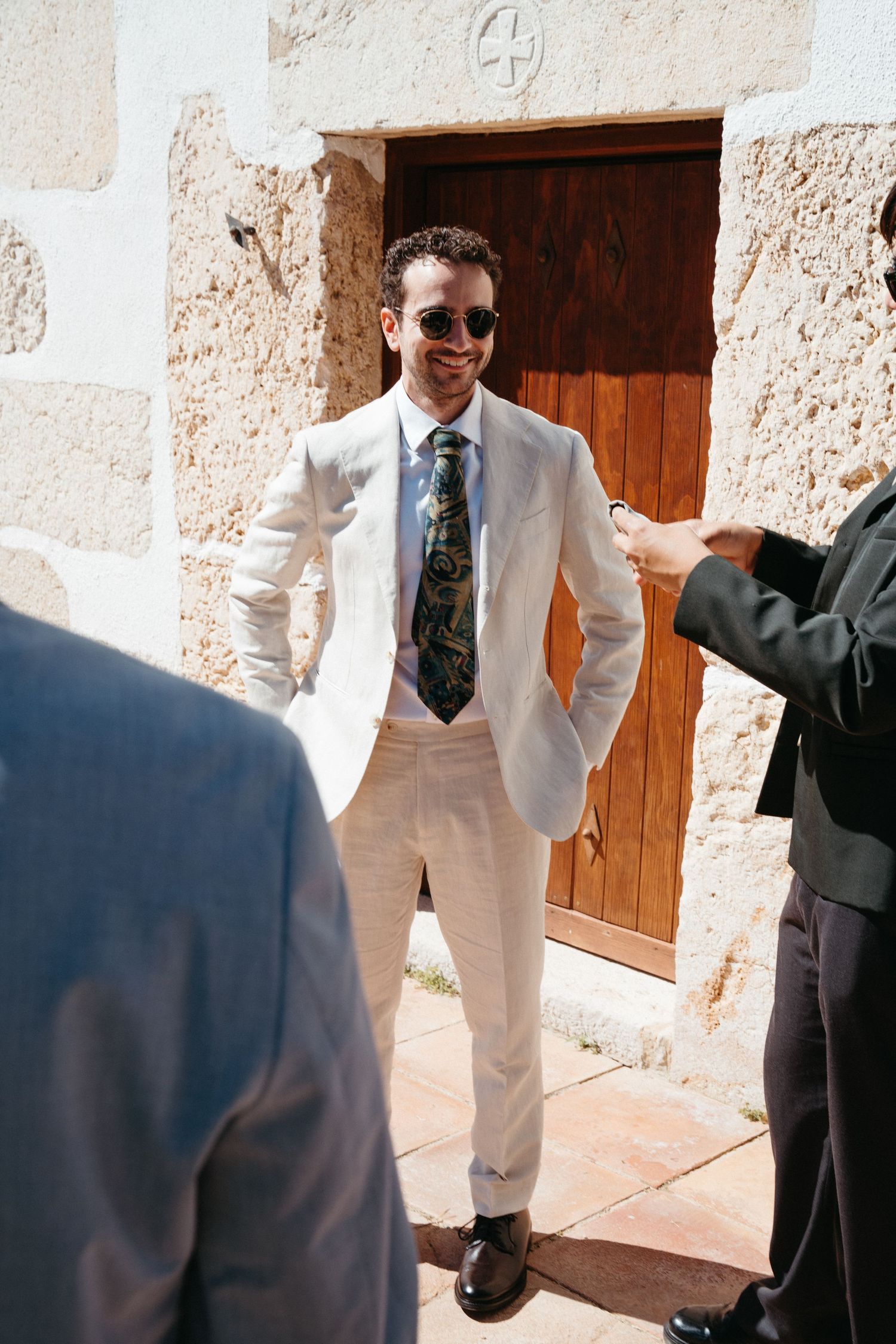 Wedding party members in suits laughing and enjoying a candid moment outside a stone building.