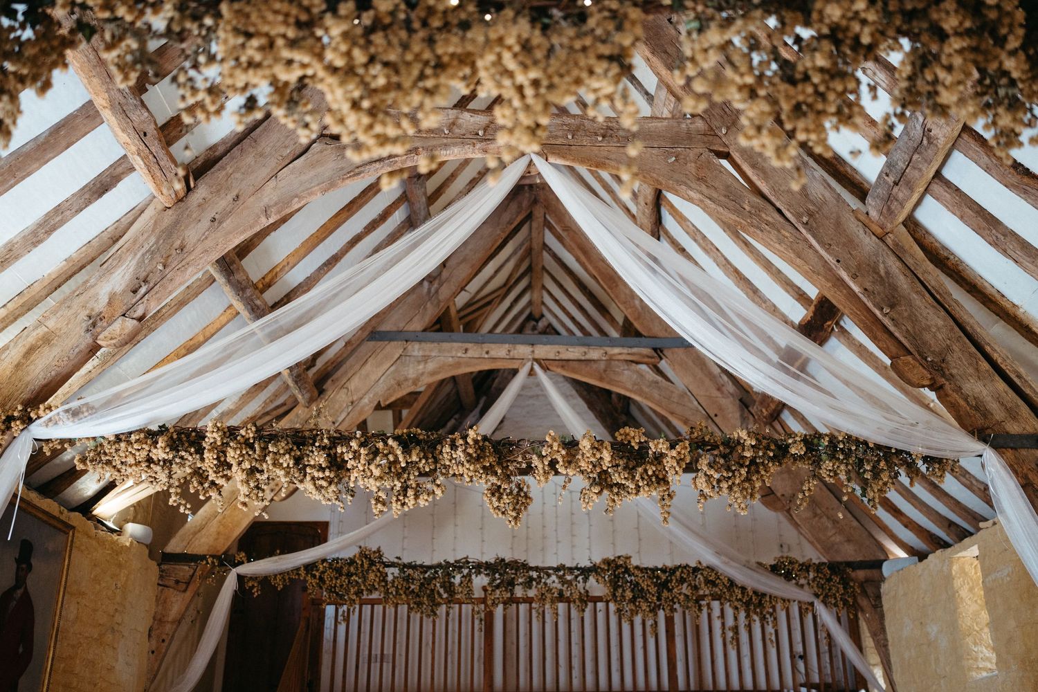Rustic wedding barn interior with white fabric drapes and autumn foliage decorating exposed wooden beams.