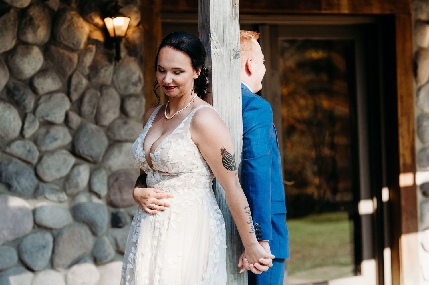 A bride in a white lace wedding dress stands holding hands with someone in a blue suit outside a stone building.