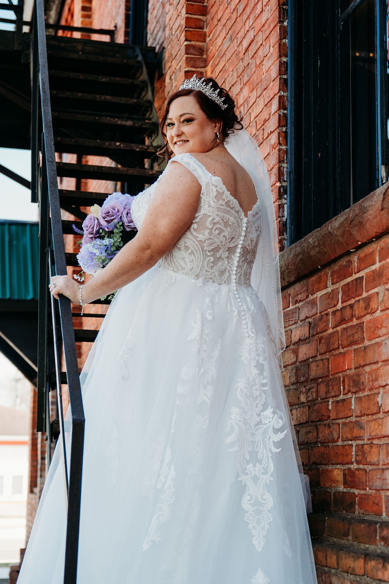 A wedding dress with intricate lace detail and flowing skirt against a rustic brick wall backdrop.