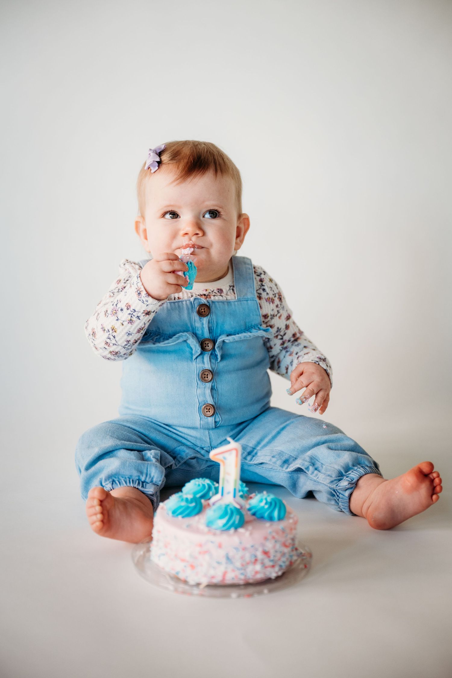 Baby in blue overalls celebrates first birthday with small decorated cake on white background.
