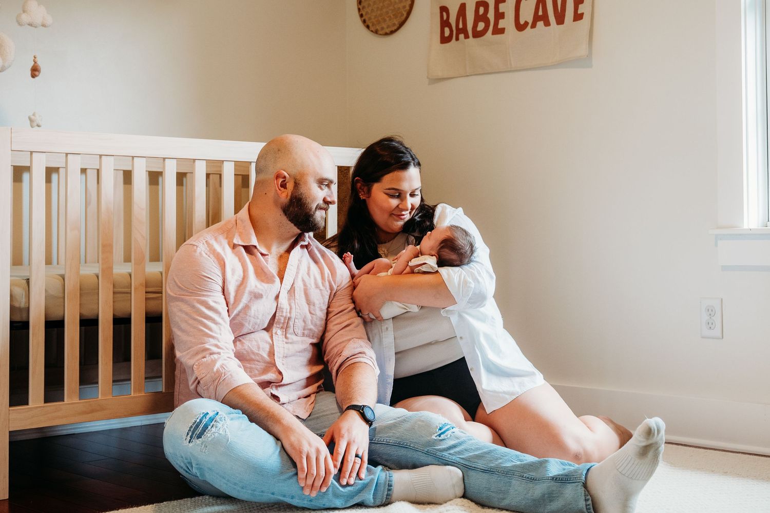 A couple sits on the floor of a nursery with their newborn baby beneath a 'Babe Cave' sign.