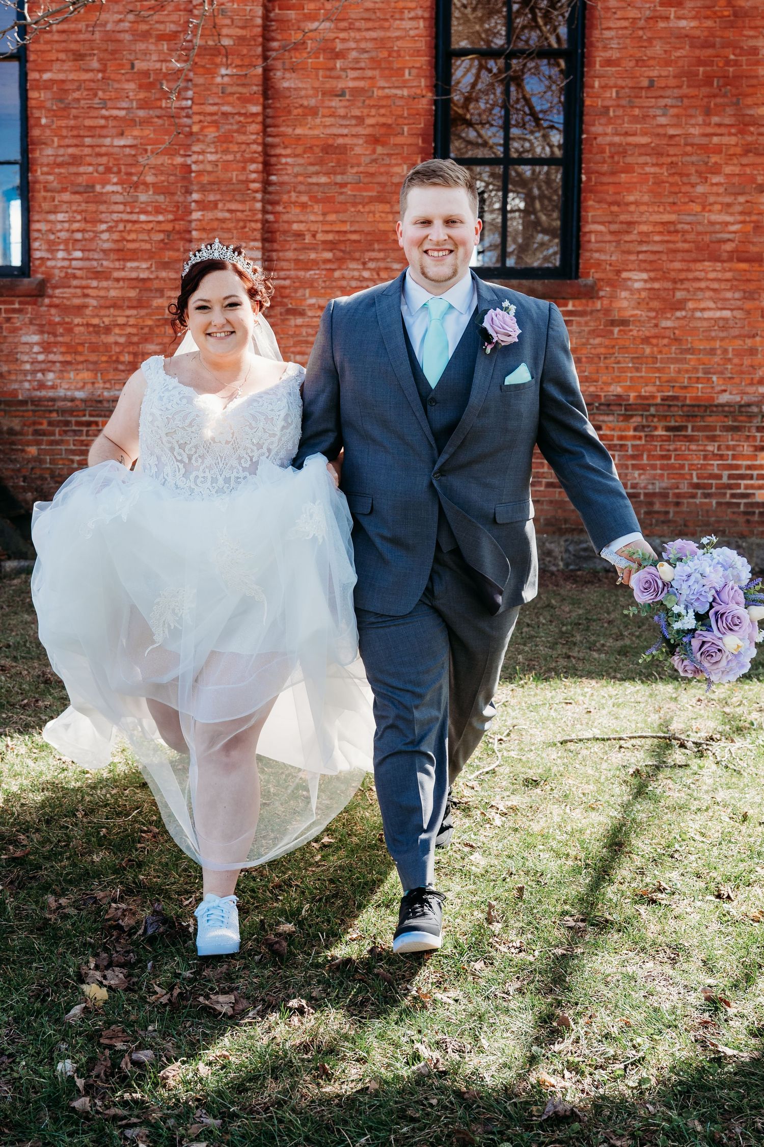 Wedding couple walking and smiling in casual sneakers against brick building backdrop.