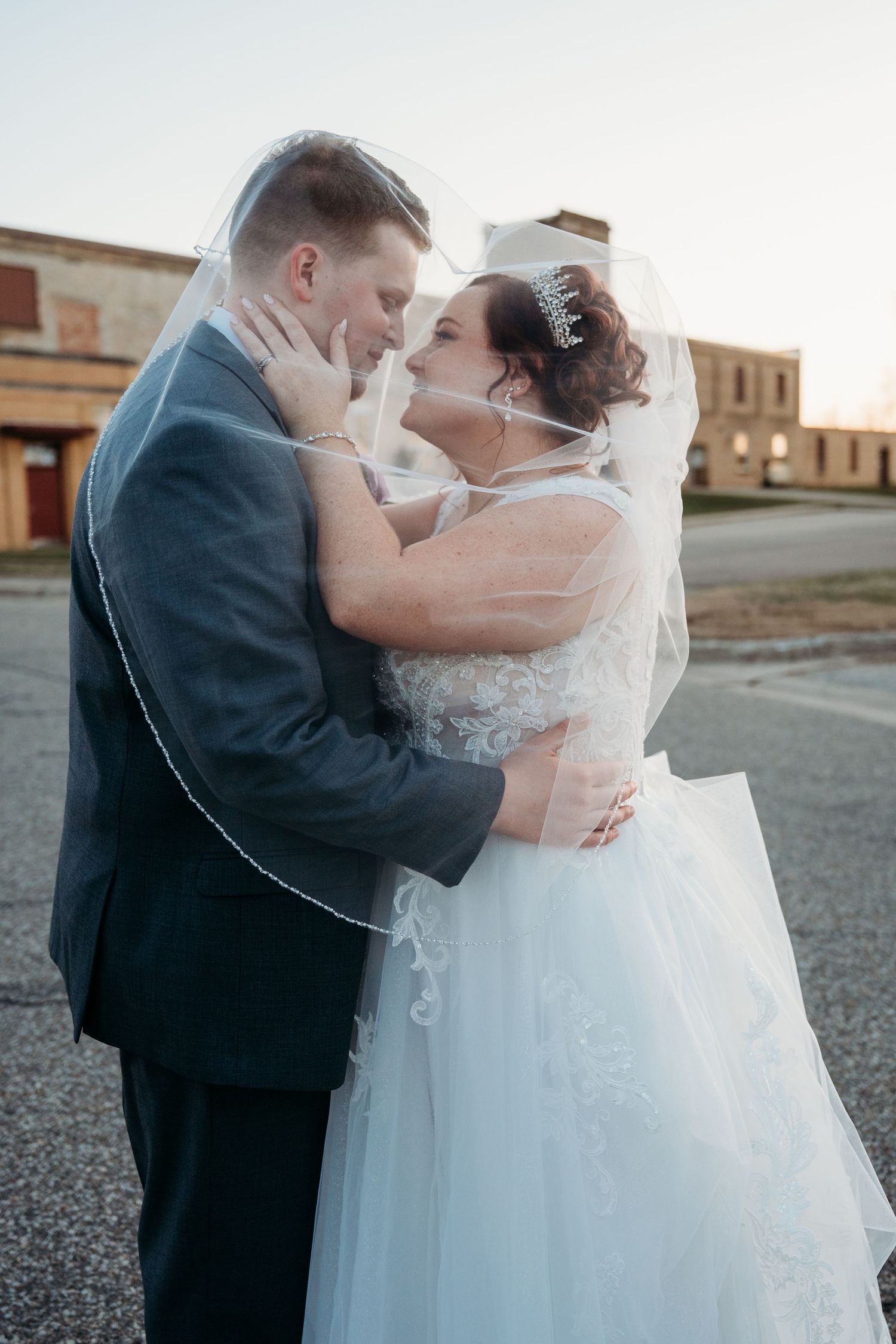A romantic wedding portrait with couple embracing outdoors at sunset near brick buildings.