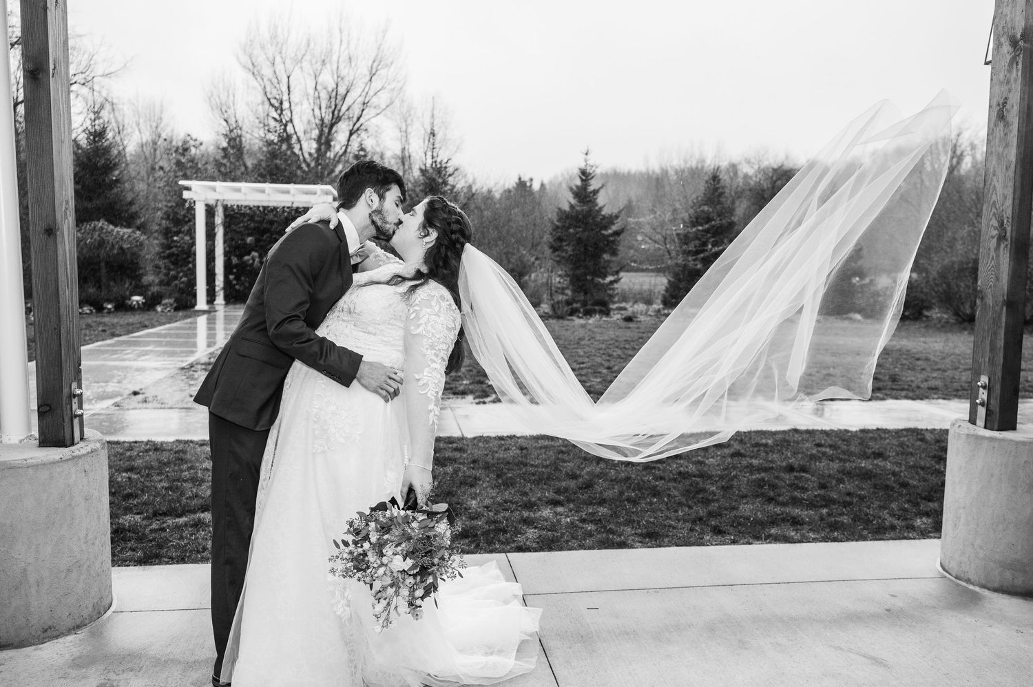 A couple shares a romantic outdoor wedding moment as the bride's veil flows dramatically in the wind at a scenic venue.