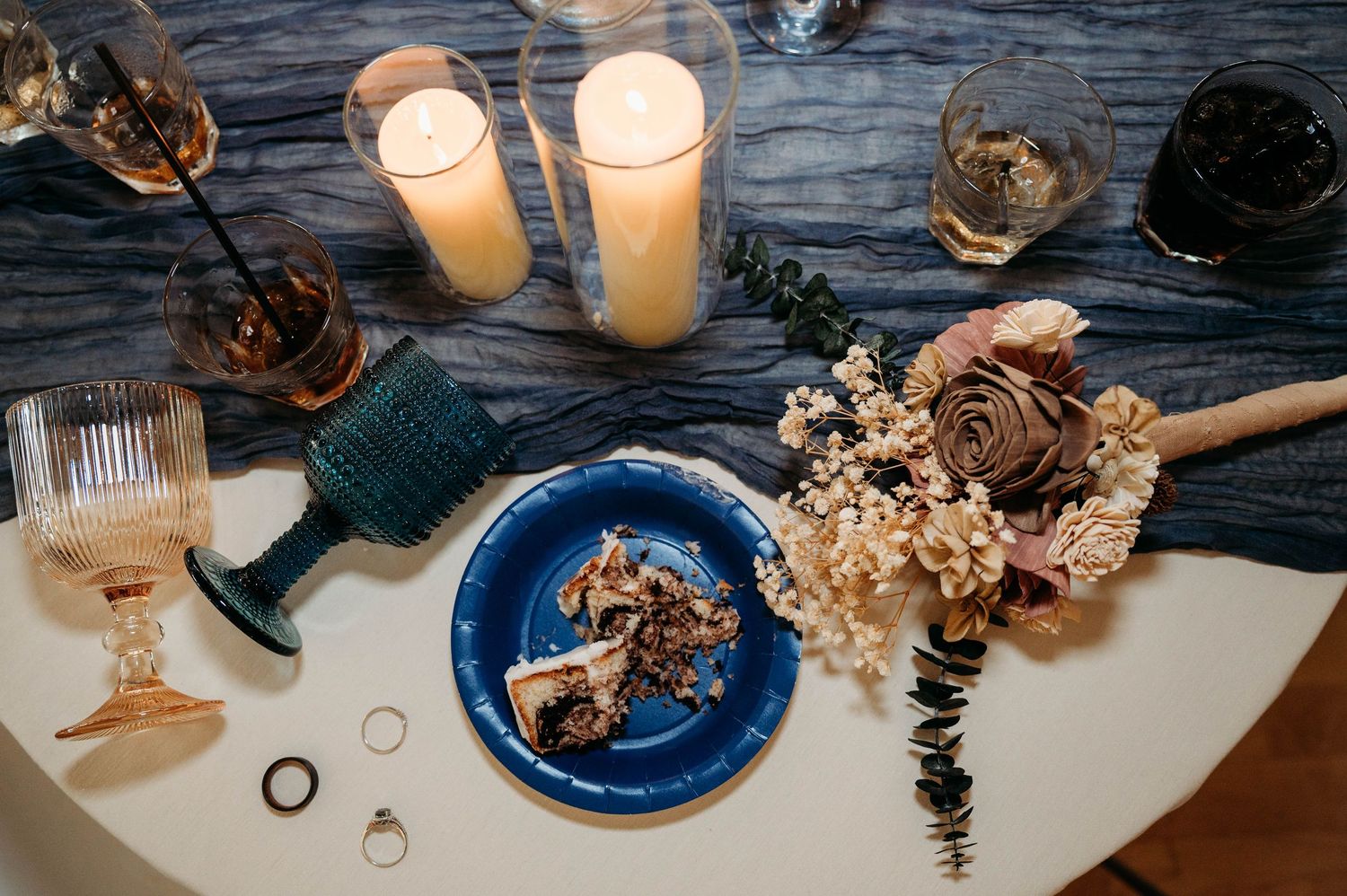 Romantic table setting with cake slice on blue plate, candles, wine glasses, and dried flowers on rustic wooden surface.