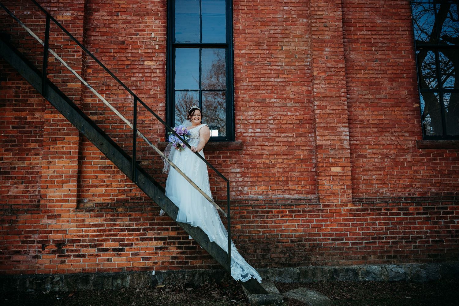 Bride in flowing white gown poses on rustic brick building staircase.
