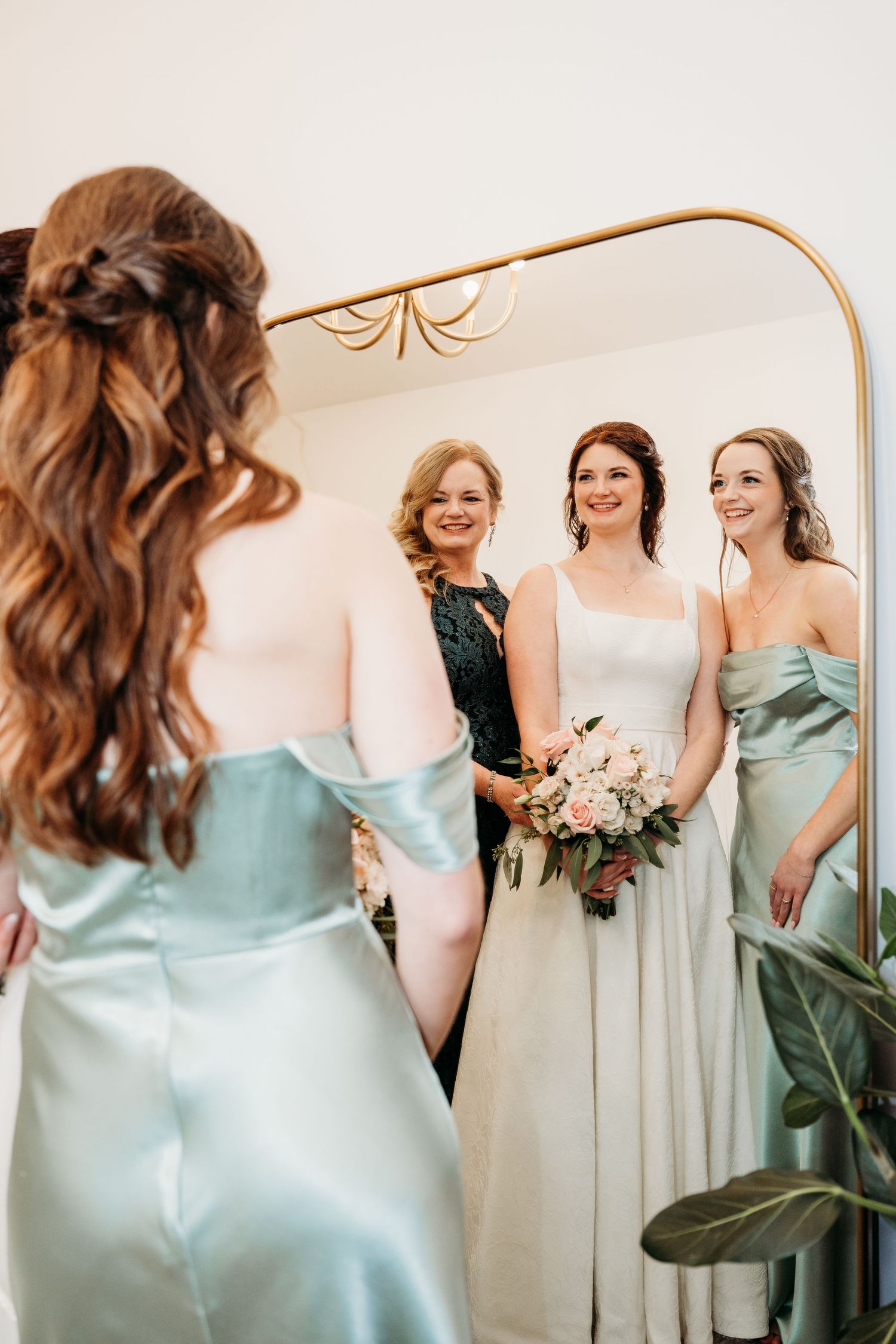 Wedding party members in mint green silk bridesmaid dresses gather around a gold-framed mirror with the bride in white.
