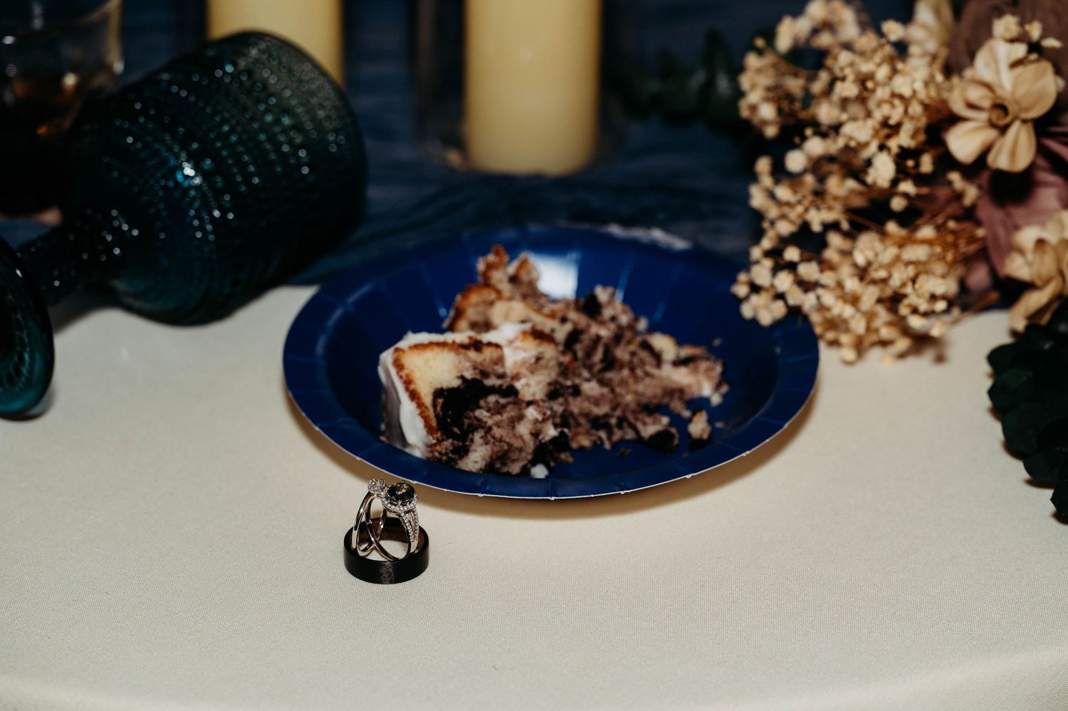 A small crystal ring displayed on a blue plate with dried flowers and a dark bottle in the background.