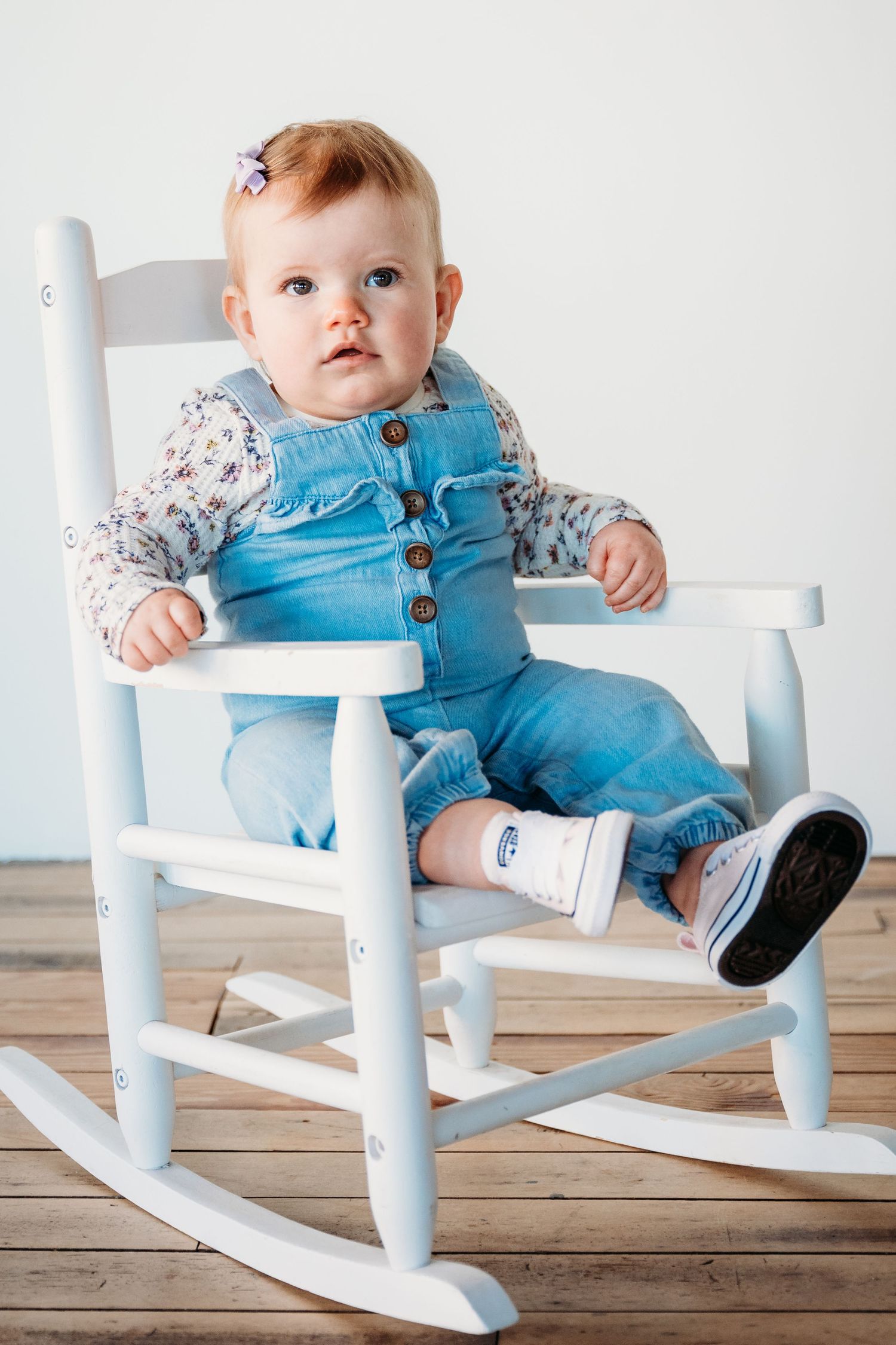 Baby in light blue overalls sitting in a white wooden rocking chair on hardwood floor, photographed from different angles.