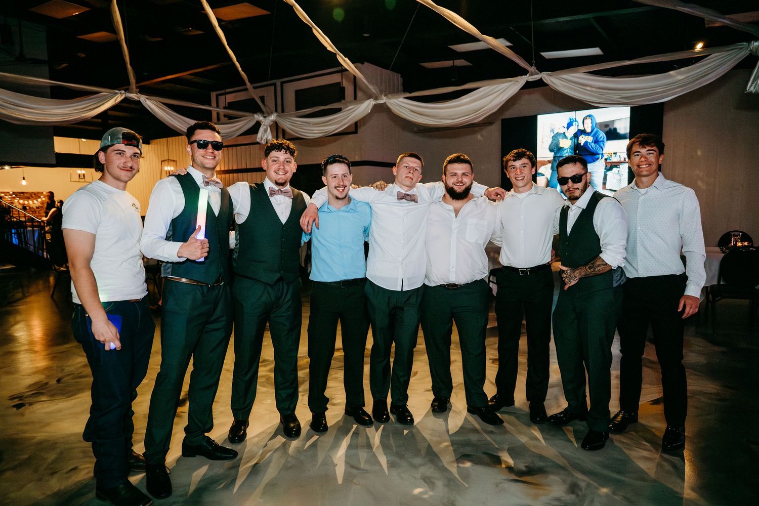 A group of groomsmen wearing green vests and matching ties stand together at a wedding reception venue.