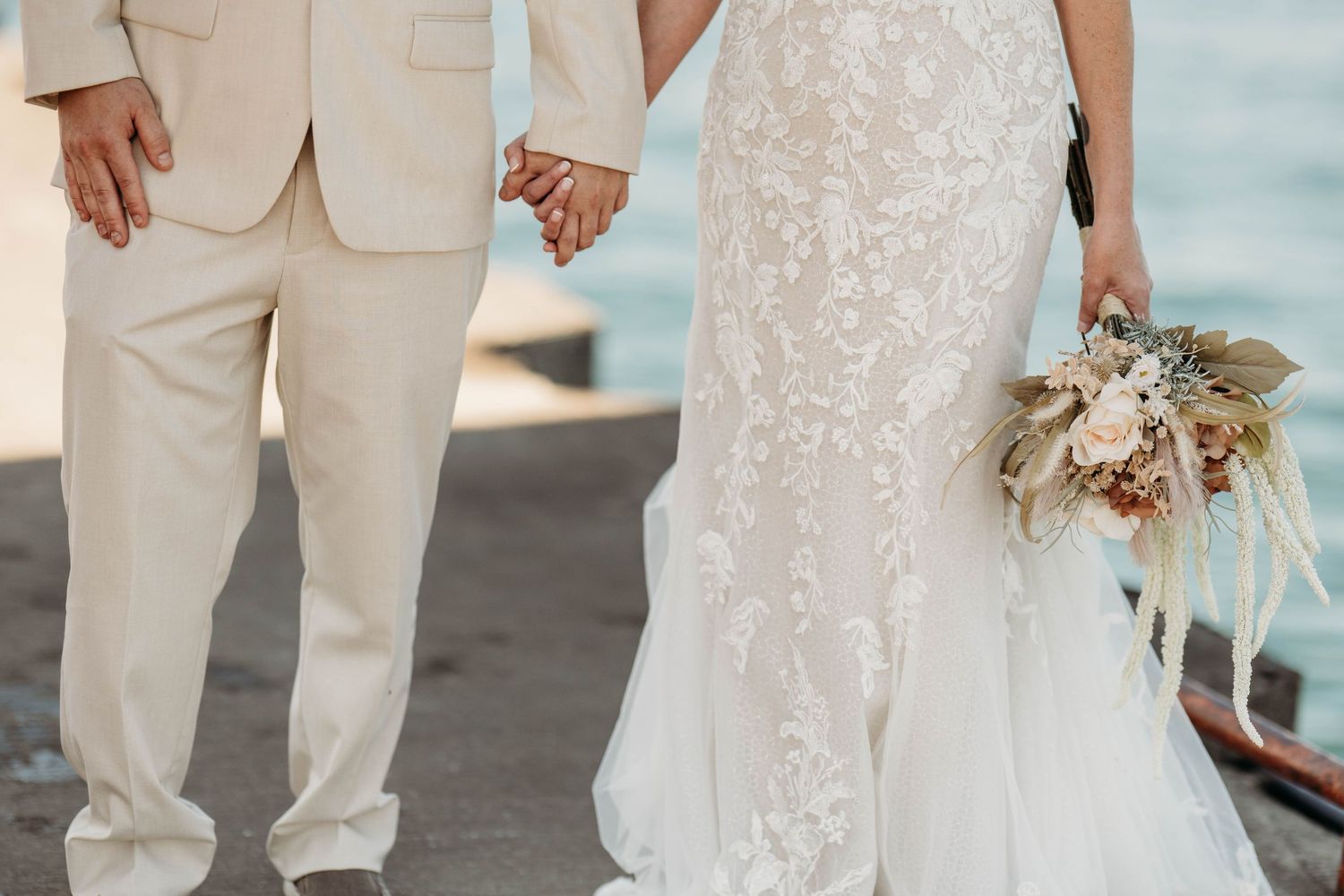A bride in a lace gown holds a dried floral bouquet next to someone wearing a beige suit at a waterfront wedding.