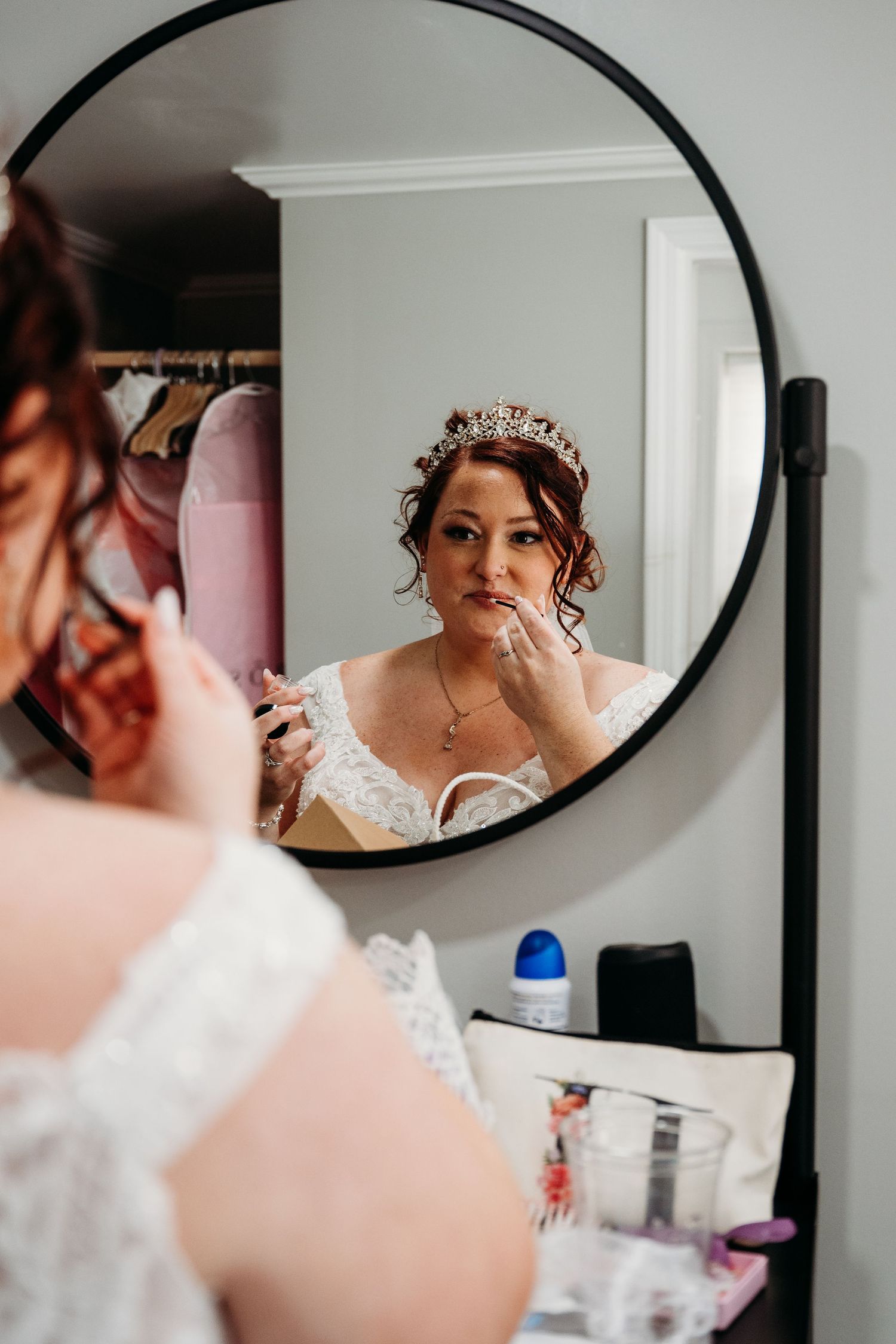 A reflection in a round black-framed mirror shows a bride wearing a delicate lace dress and ornate gold crown.