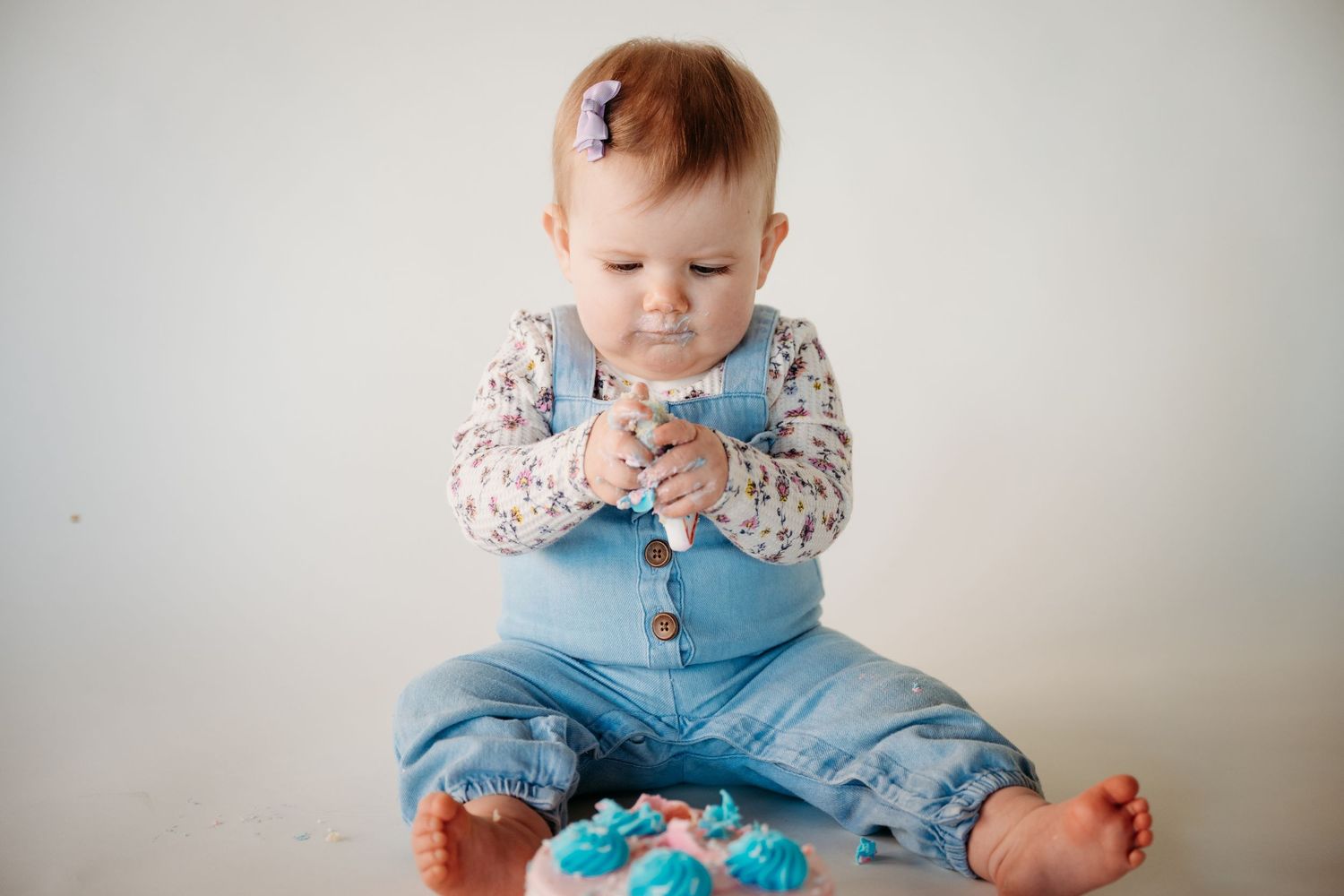 Baby wearing floral shirt and blue overalls sits on white floor playing with a turquoise toy.