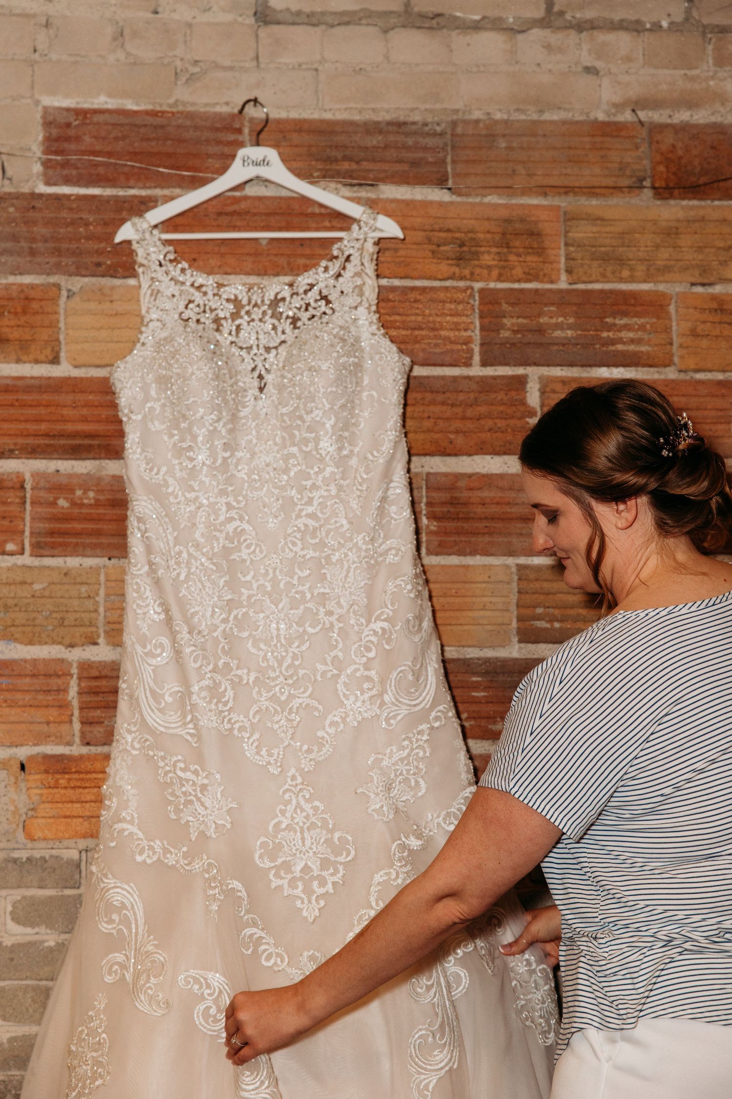 A detailed lace wedding dress hangs on a white hanger against a brick wall while being prepped for a special occasion.