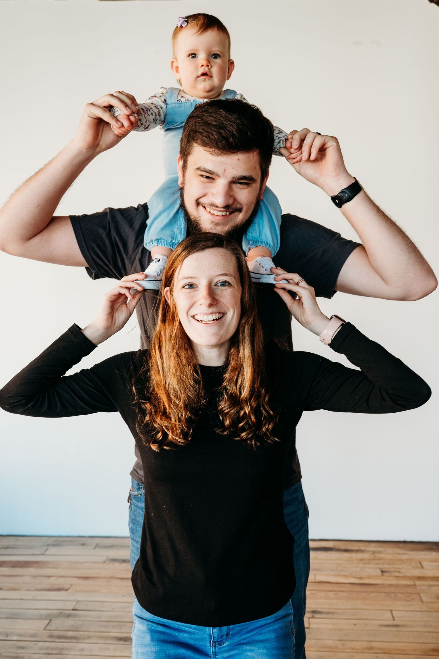 A happy family poses together with a baby sitting on shoulders against a white wall background.
