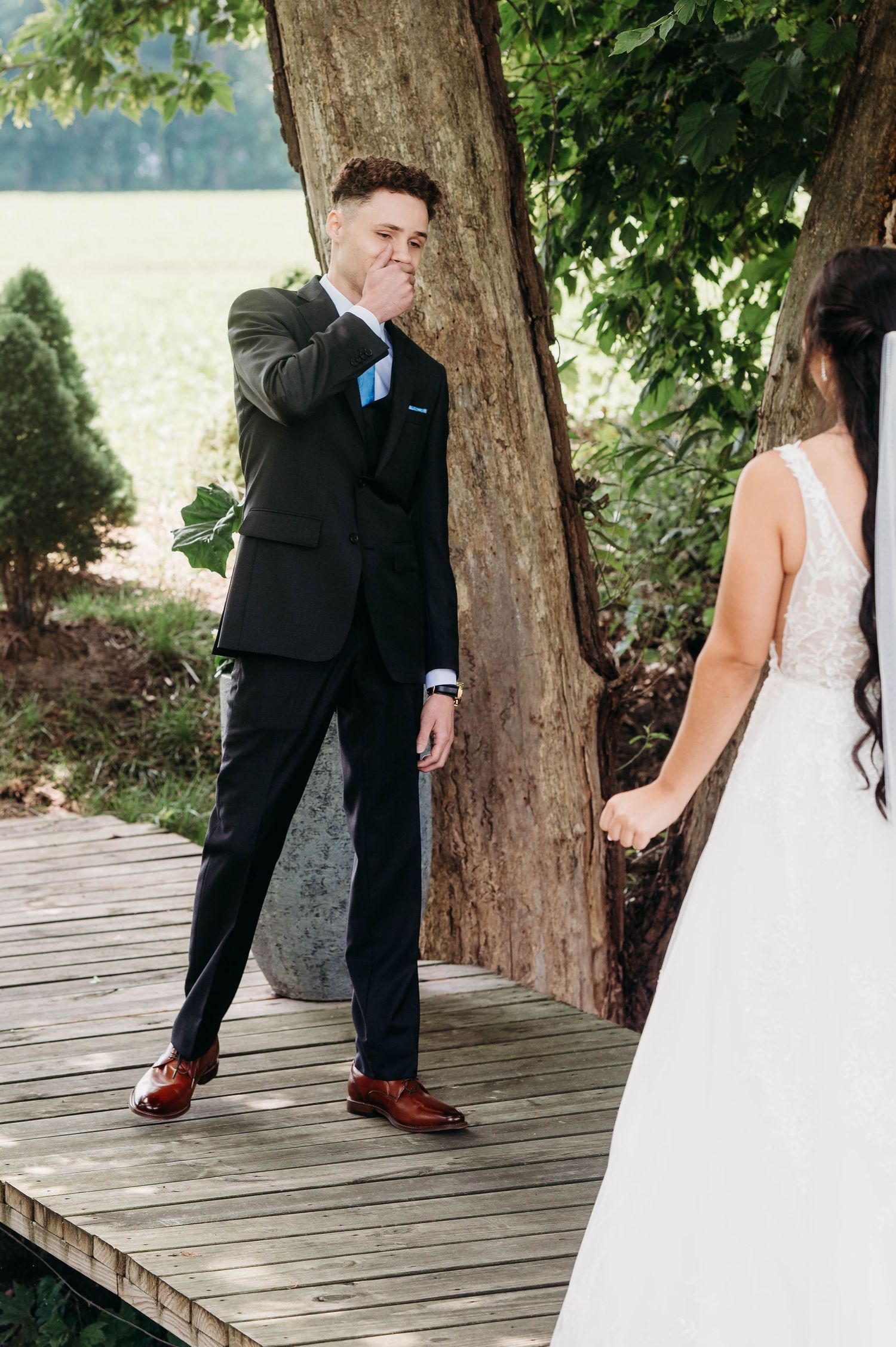 An emotional first look moment between groom in black suit and bride in white dress on wooden dock by the water.