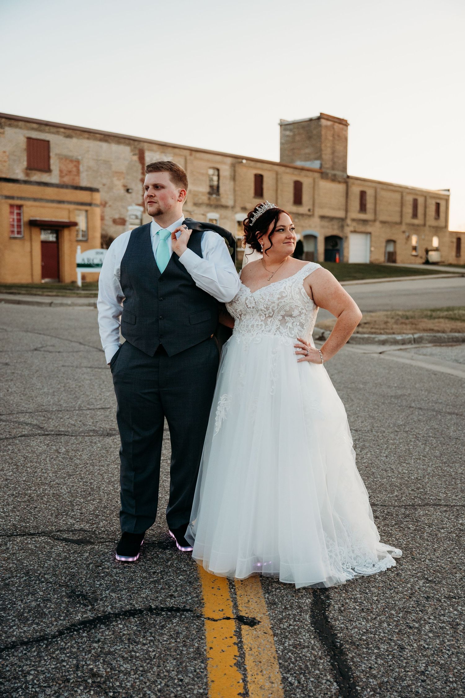 A bride and groom stand together on a street at sunset, posing in front of an industrial brick building.