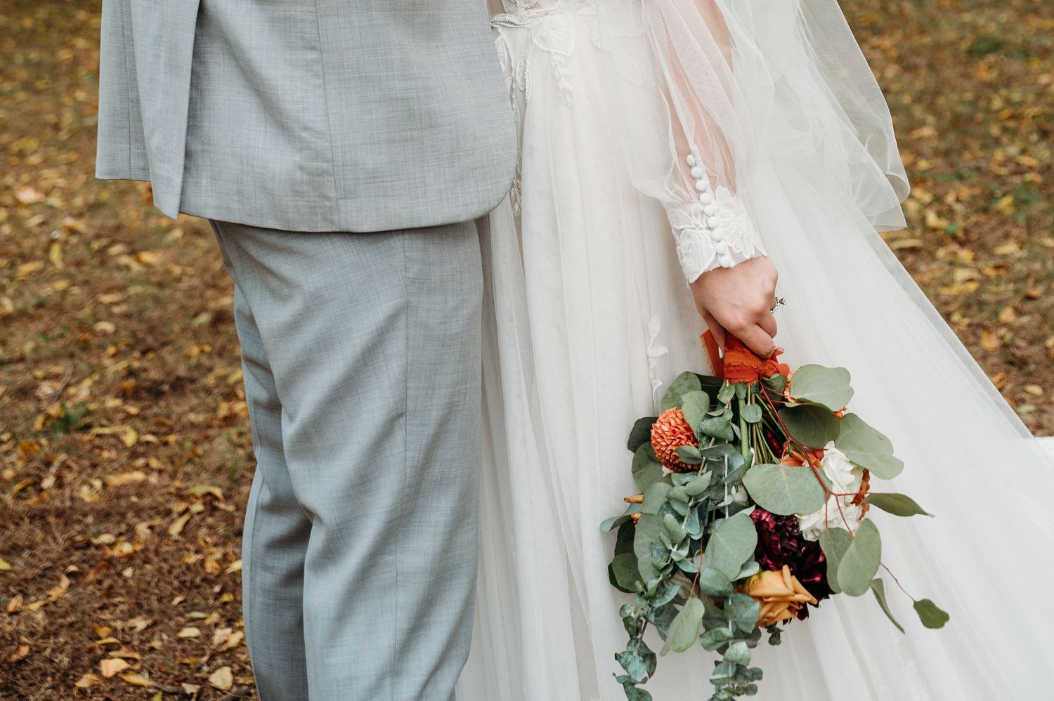 Wedding couple in grey suit and white dress holding floral bouquet with eucalyptus leaves on autumn ground.
