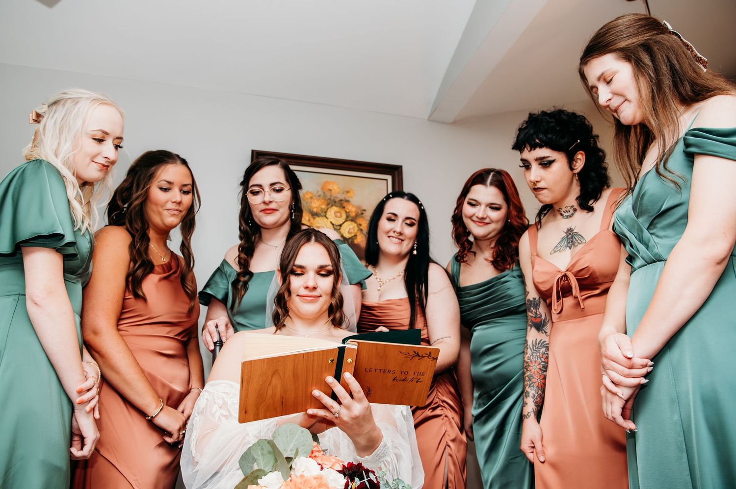 A group of bridesmaids wearing coral and emerald dresses gather around looking at a book during wedding preparations.
