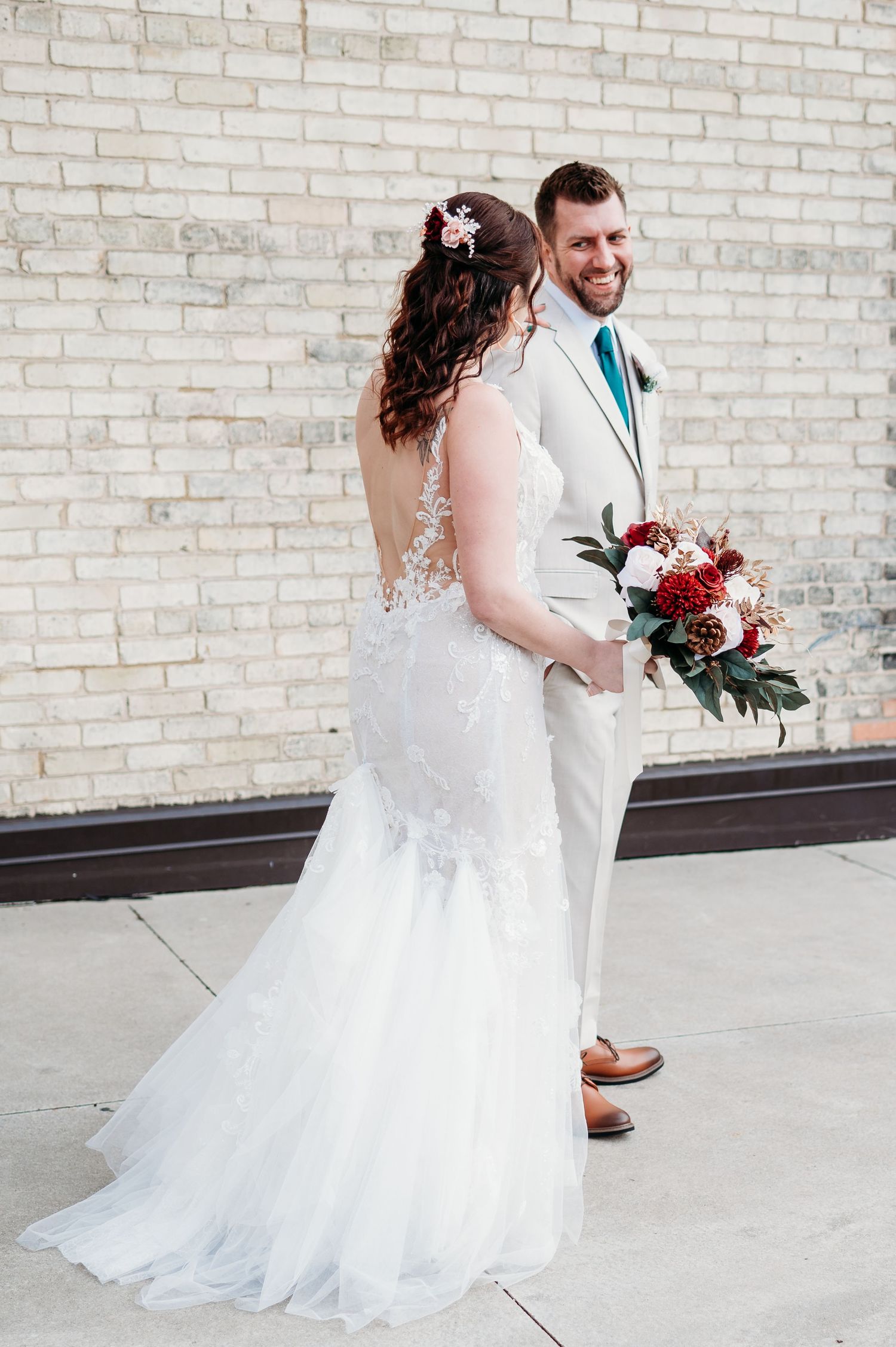 A bride in a white lace gown holding a red bouquet smiles at her partner against a brick wall.