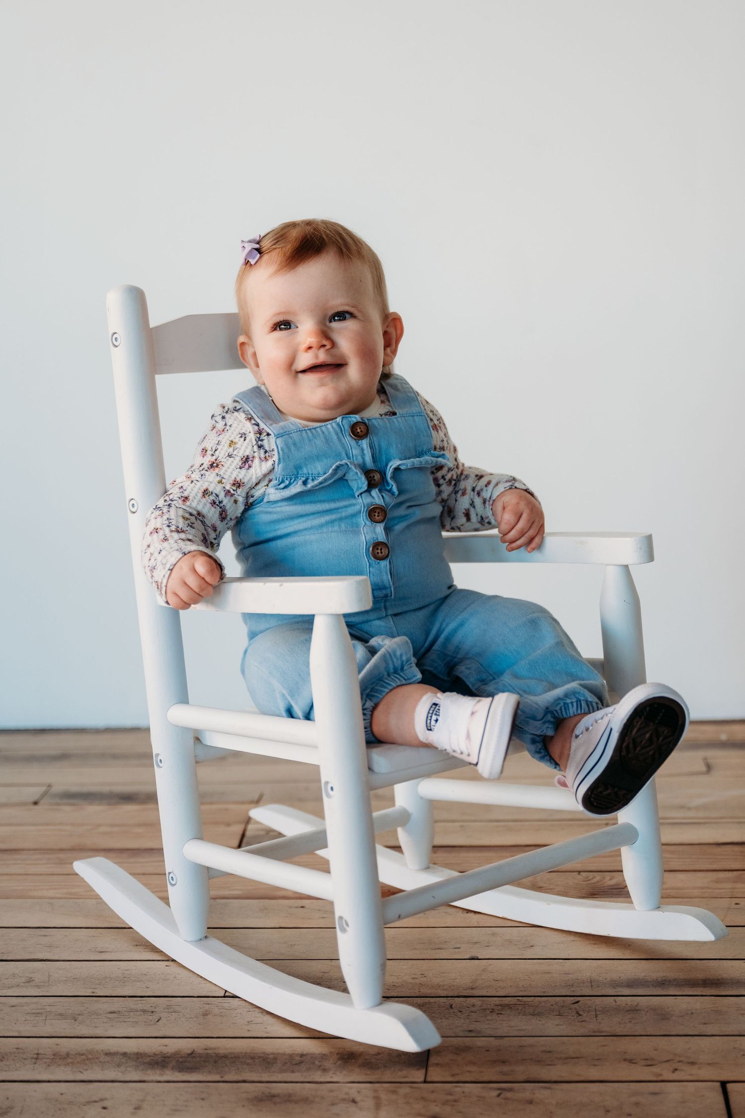 A small child wearing blue overalls sits and smiles in a white wooden rocking chair on hardwood flooring.