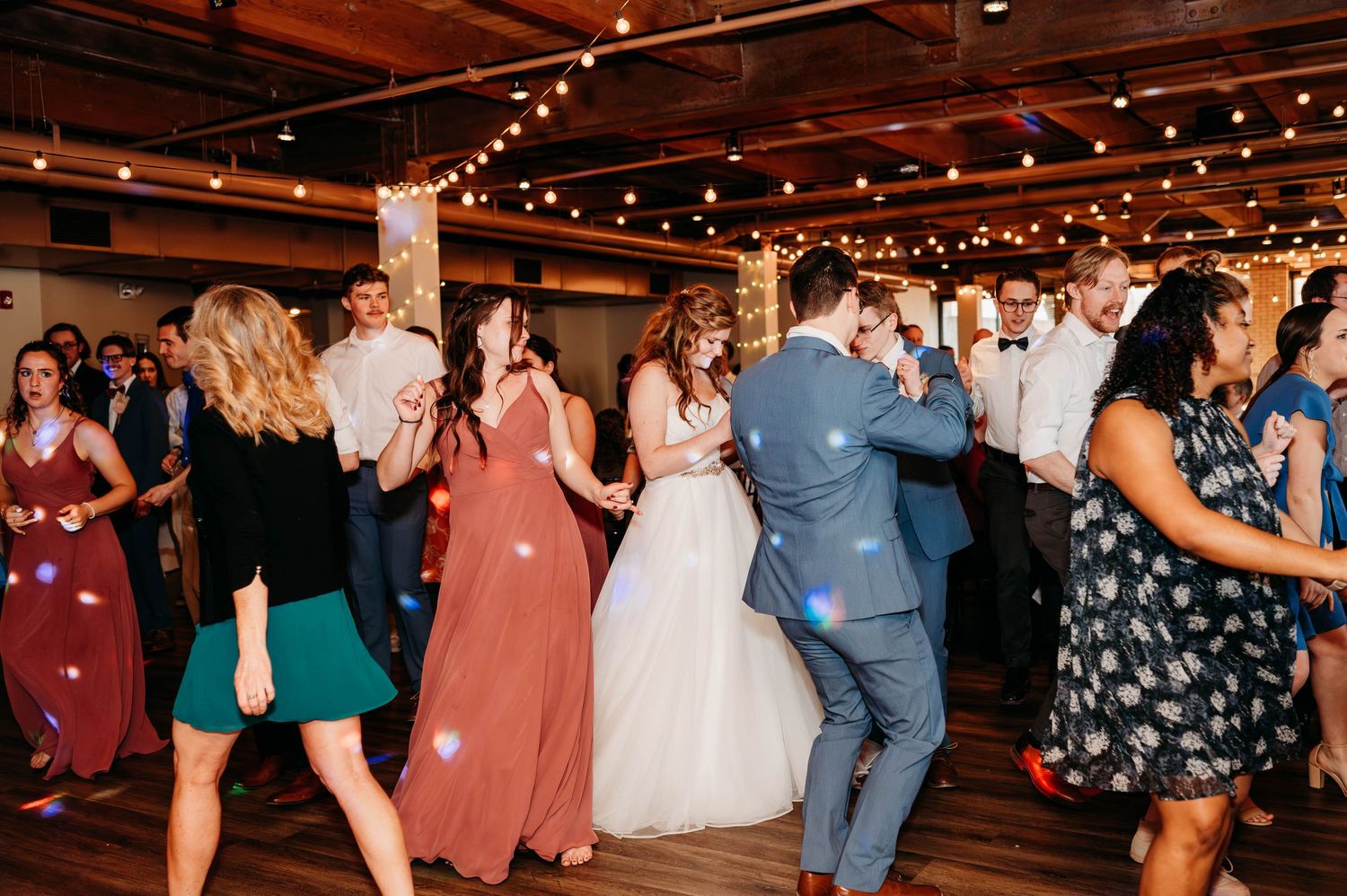 Wedding guests dance and celebrate on a rustic wooden dance floor under warm string lights at an indoor reception.