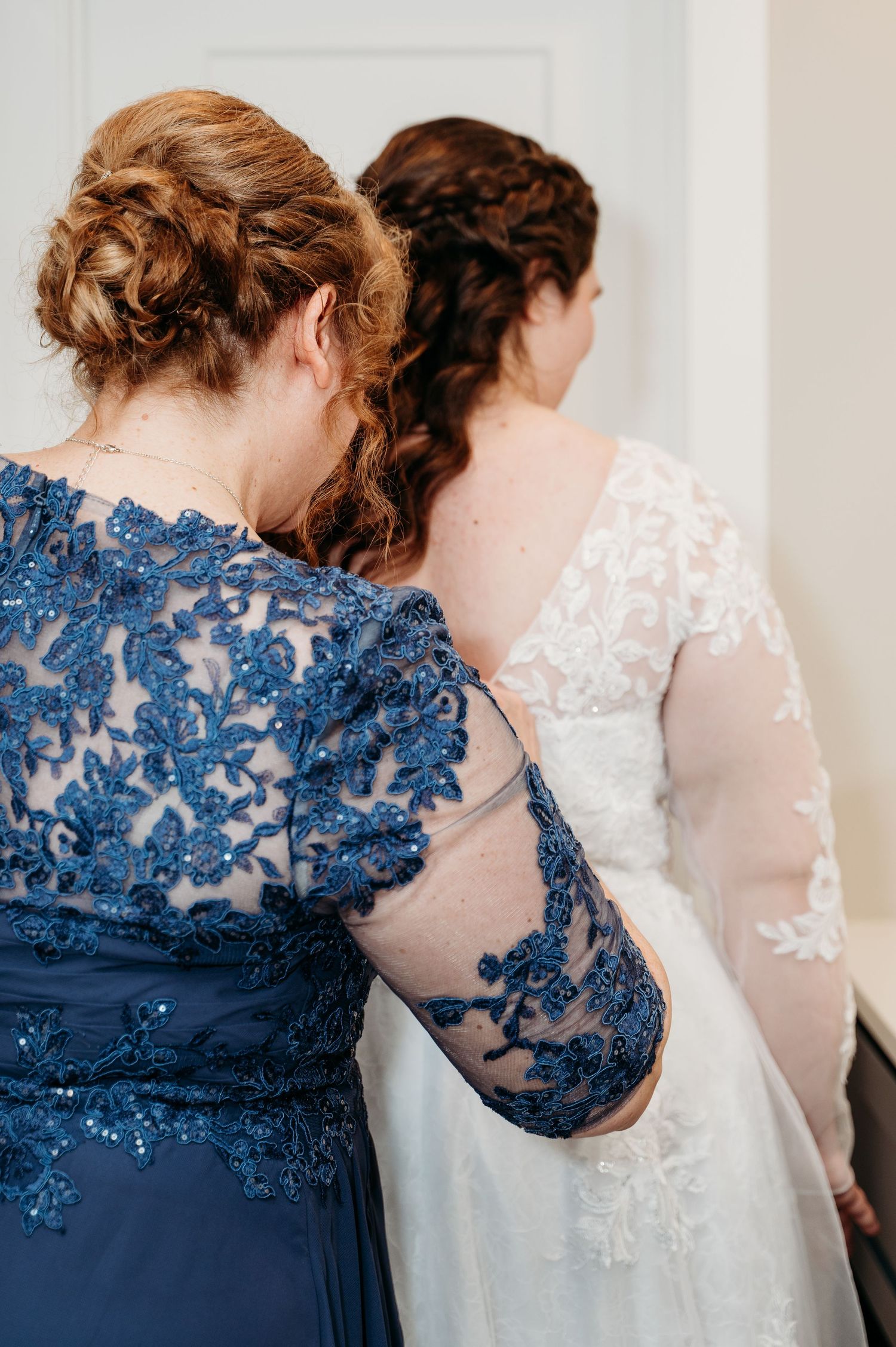 Two women wearing elegant lace dresses in navy blue and white stand back to back with upswept hairstyles.