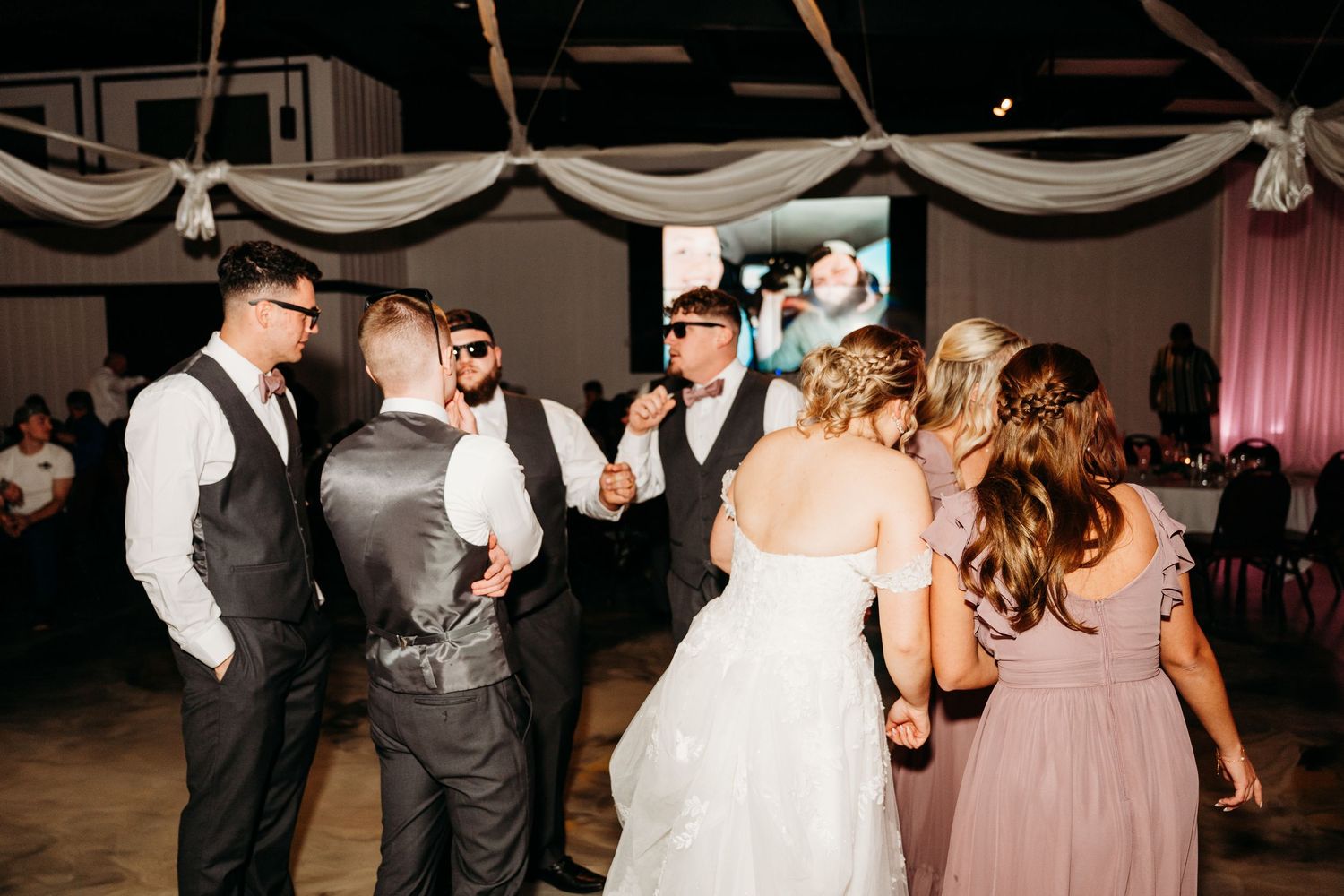 Wedding guests dance and celebrate together on a reception dance floor with white draping decor overhead.