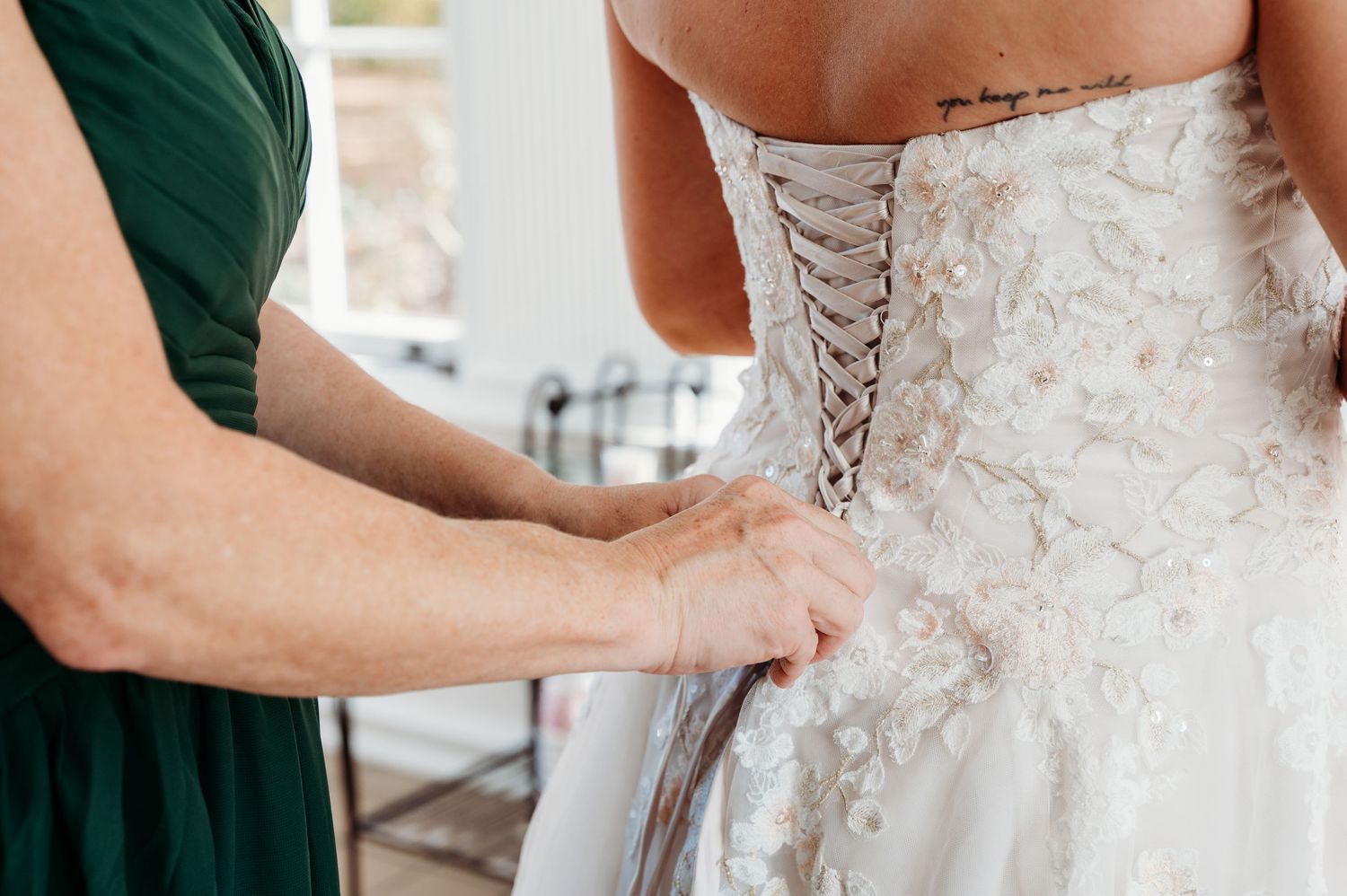 A bridesmaid in emerald green helps lace up the back of an ornate white lace wedding gown.
