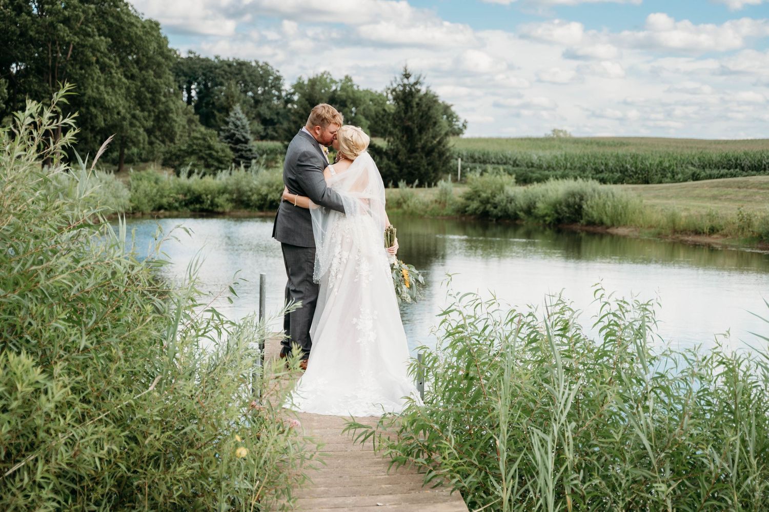 A couple shares a romantic moment on a wooden dock overlooking a serene pond surrounded by tall grass and farmland.