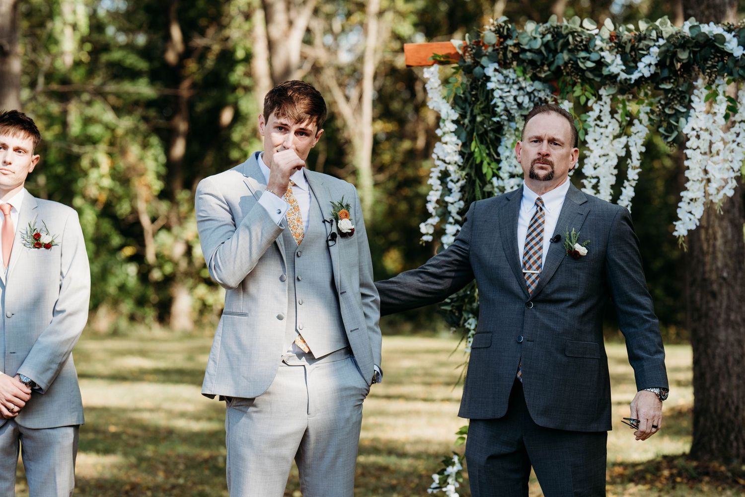 A joyful outdoor wedding celebration with groomsmen in light gray suits and navy attire under a floral arch.