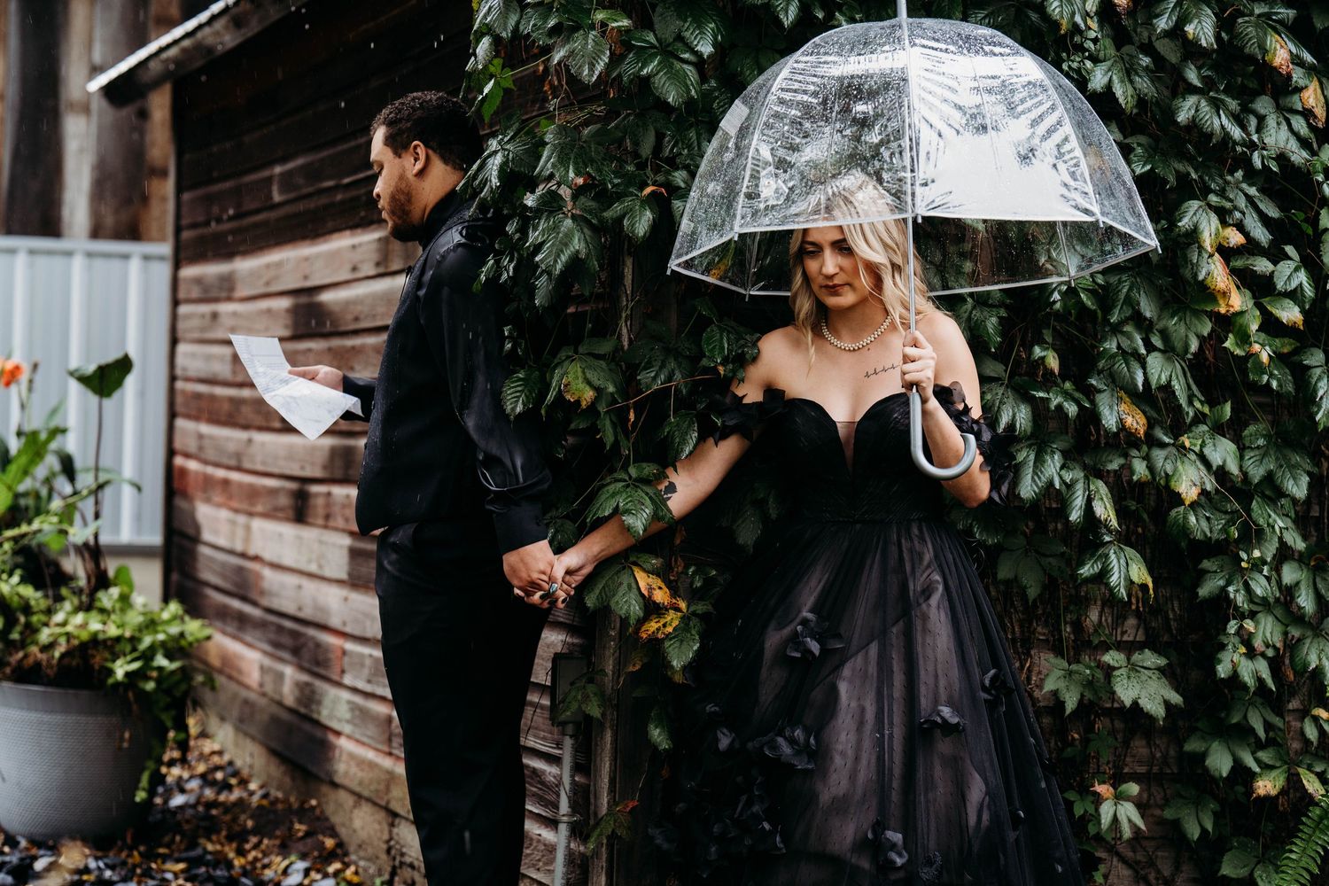 Bride in black dress holds clear umbrella while standing against ivy-covered wall during rainy day wedding.