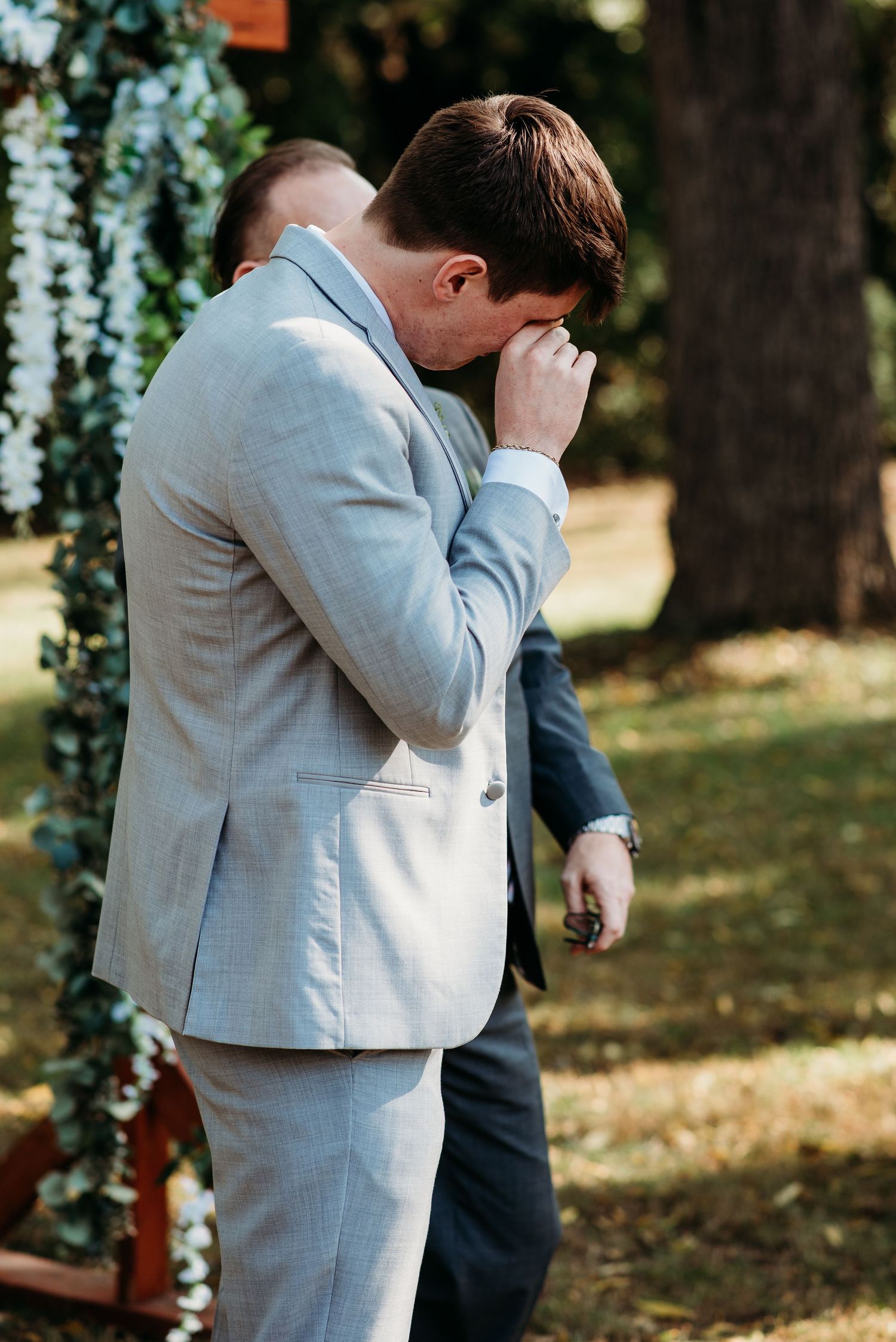 An emotional moment at an outdoor wedding ceremony as someone in a light gray suit covers their face.