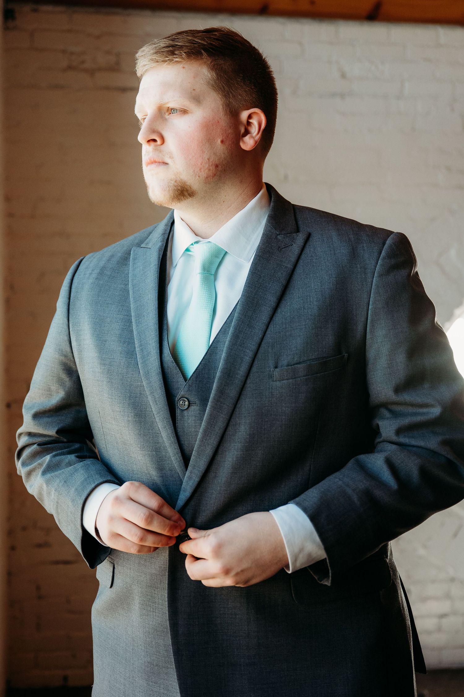A person in a grey three-piece suit adjusts their jacket while wearing a light turquoise tie.