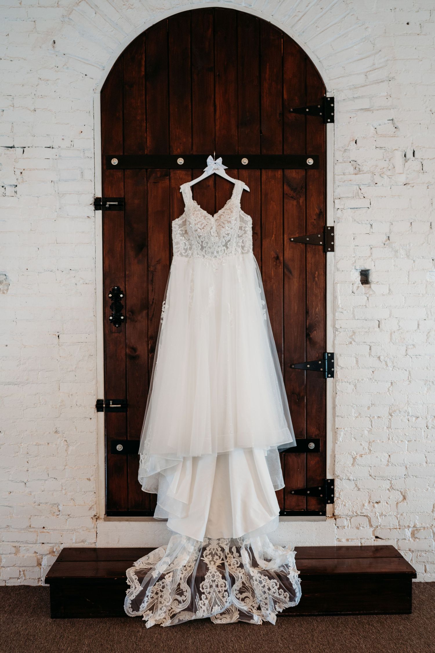 A white lace wedding dress hangs elegantly on a rustic wooden door with iron hinges against a white brick wall.