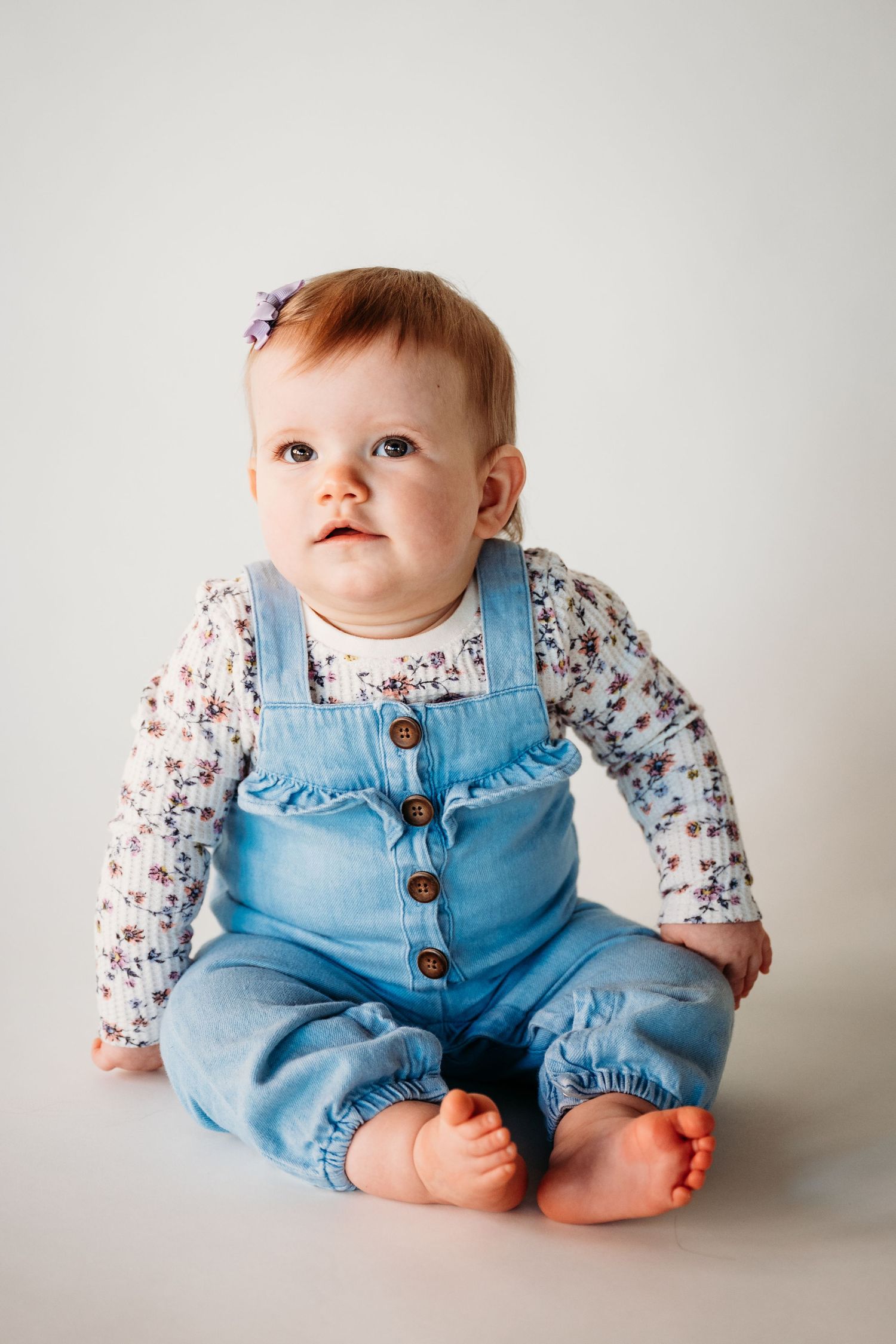 Baby in light blue denim overalls and floral print shirt sits against white background.