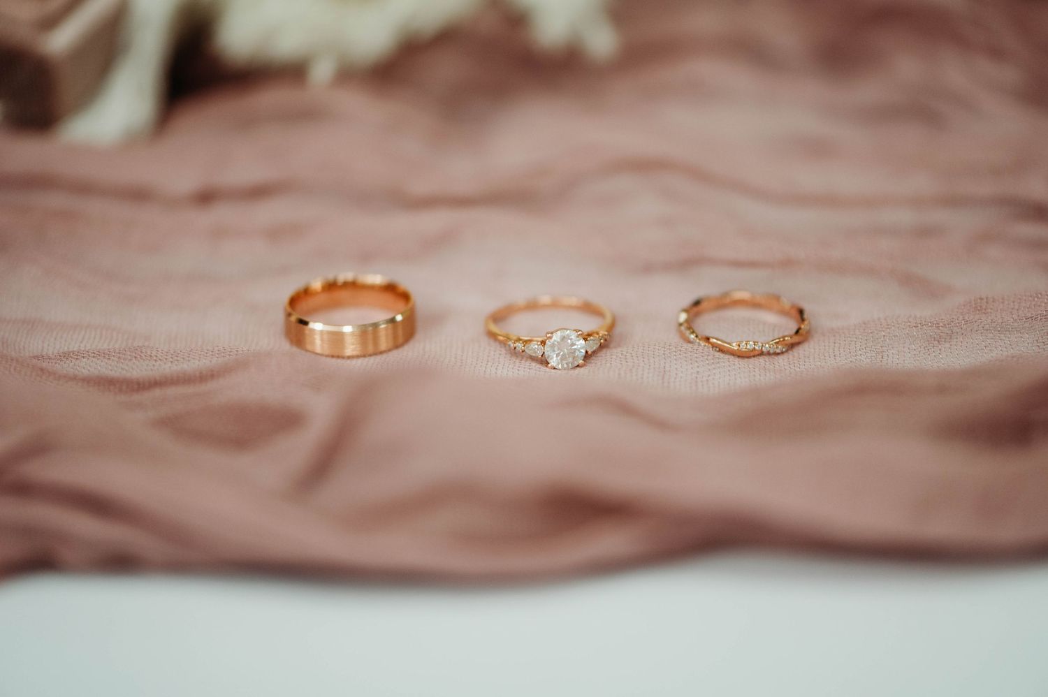 Three delicate rose gold wedding rings displayed on soft pink silky fabric with intricate designs and stones.