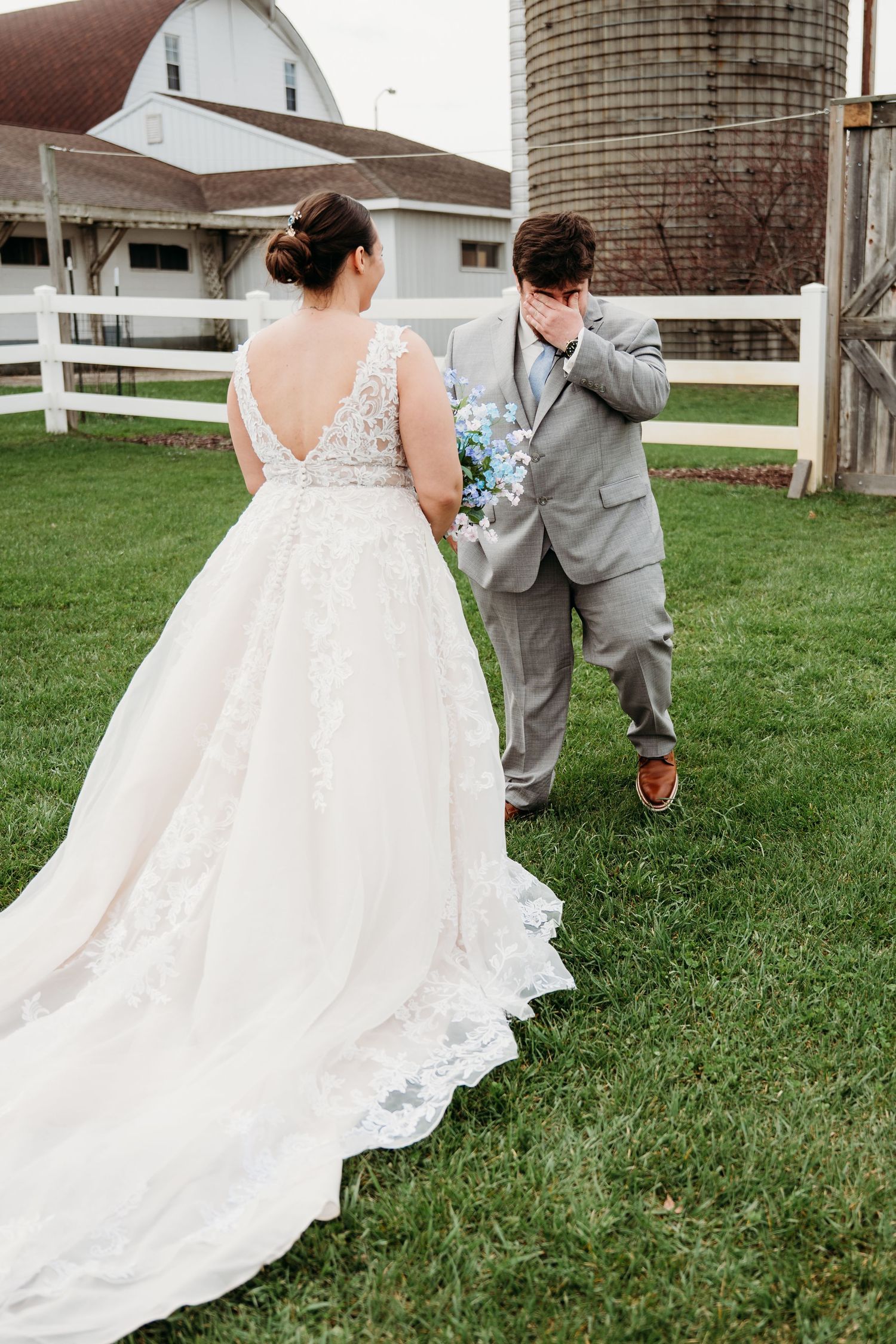 A couple in wedding attire walk together in front of a rustic white barn and silo on a grassy lawn.